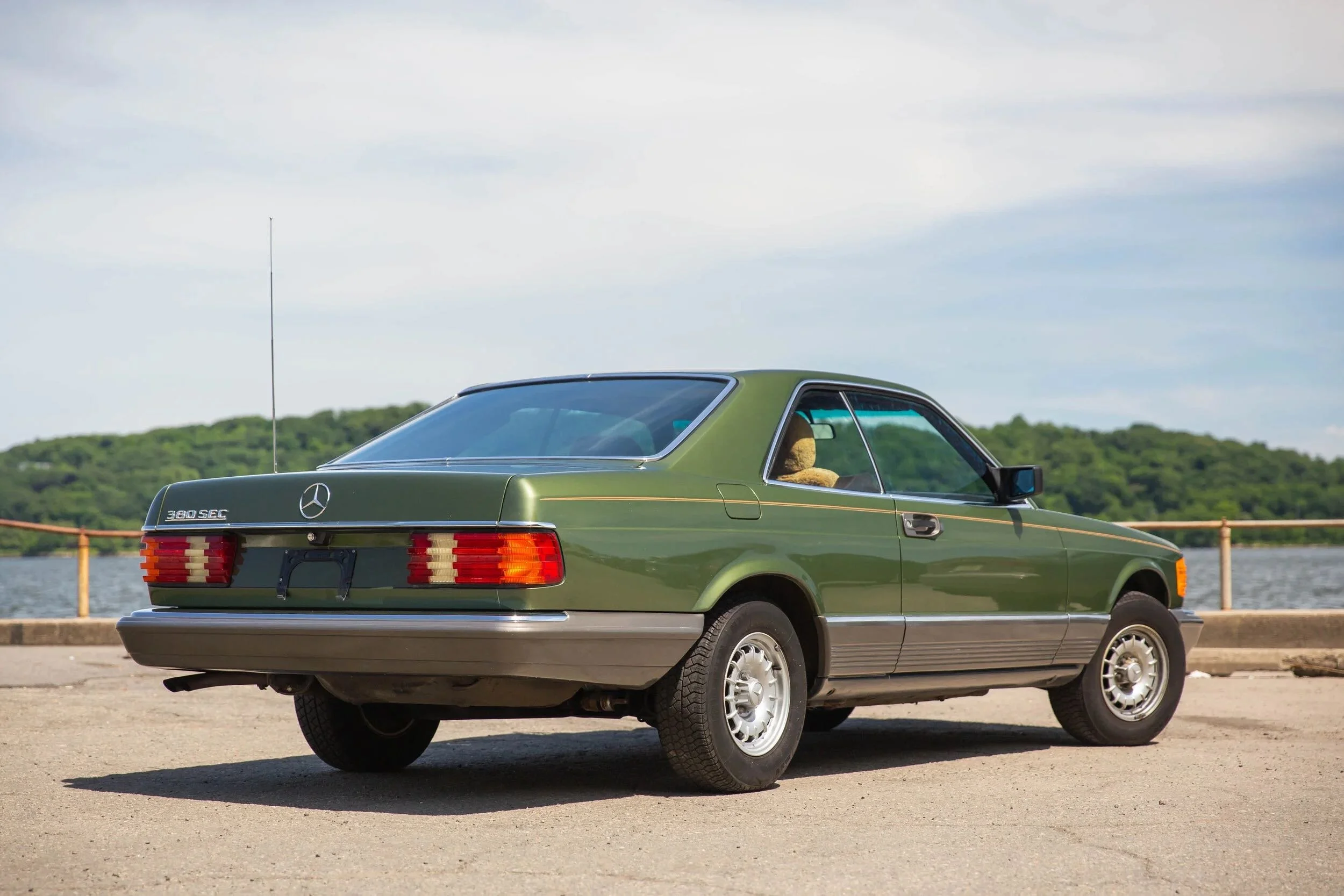 A vintage green Mercedes-Benz 380 SEC car parked near a body of water with green hills in the background.