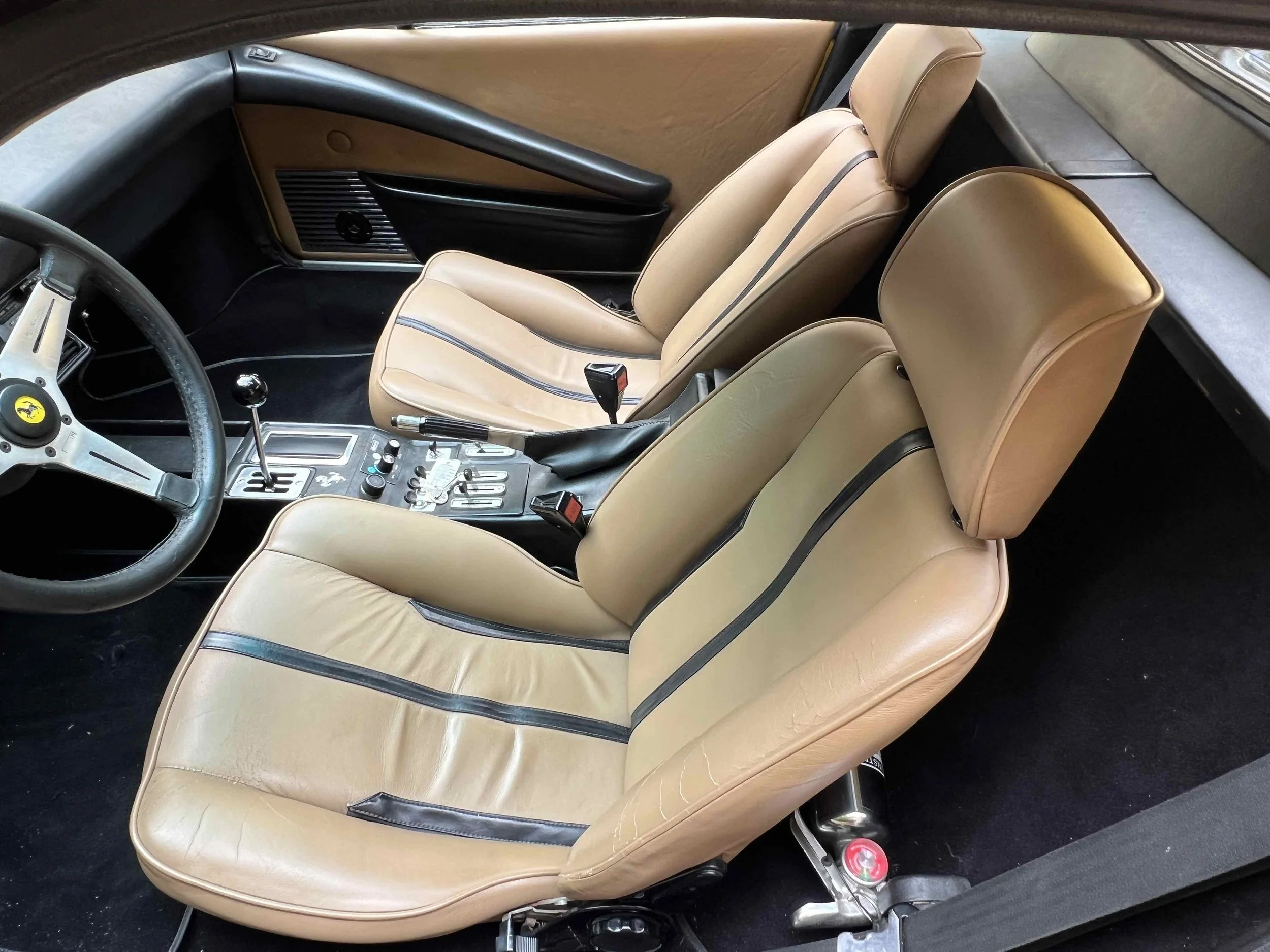 Interior of a vintage Ferrari car, showing two tan leather racing seats, a black steering wheel with Ferrari emblem, and center console with gear shifter and controls.