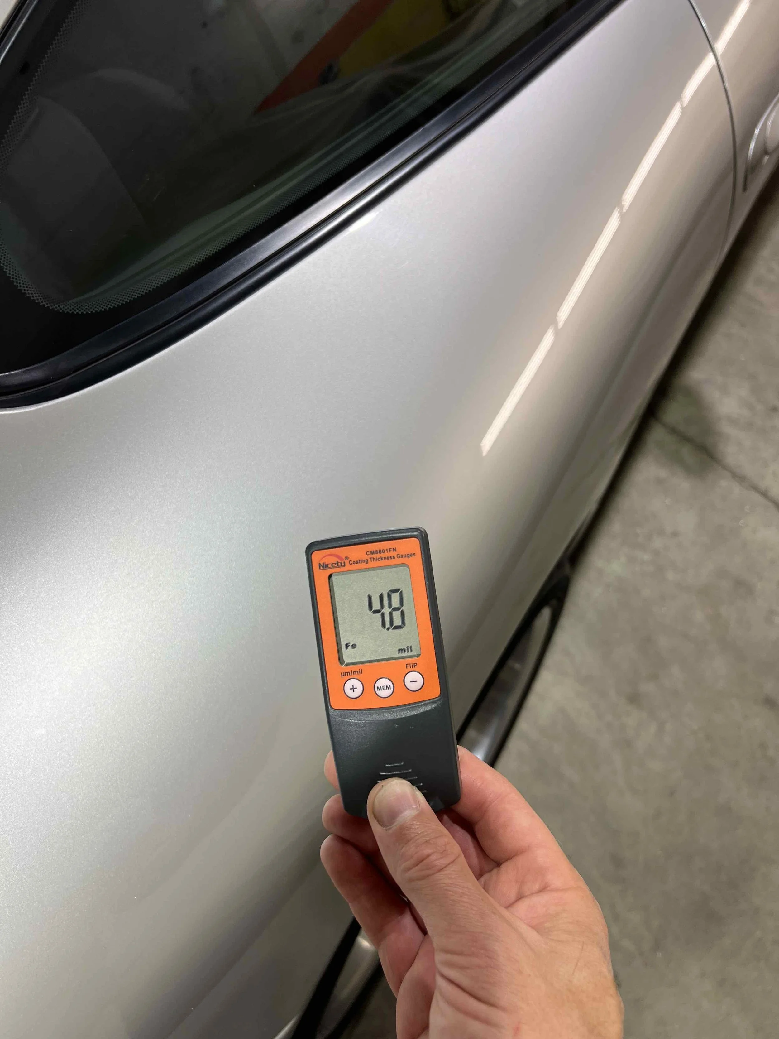 A person holding a coating thickness gauge near the side of a silver car, measuring 48 micrometers.