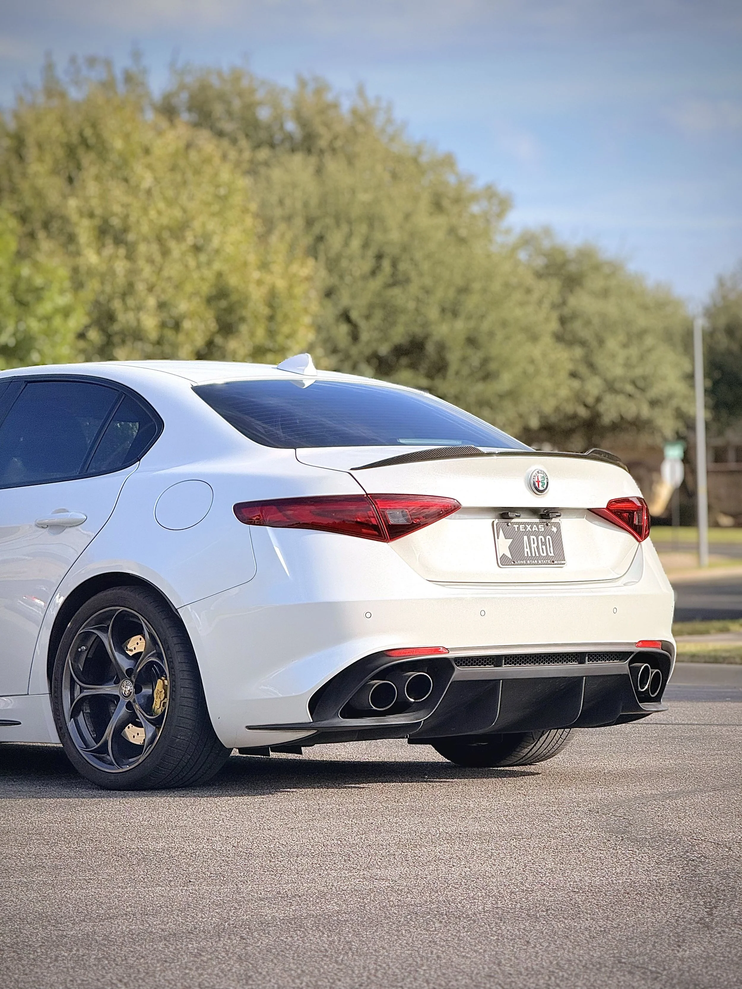 White Alfa Romeo sports car with black wheels and quad exhaust pipes parked on the road, showing the rear view with a Texas license plate reading "ARGI." Trees and a blue sky are in the background.
