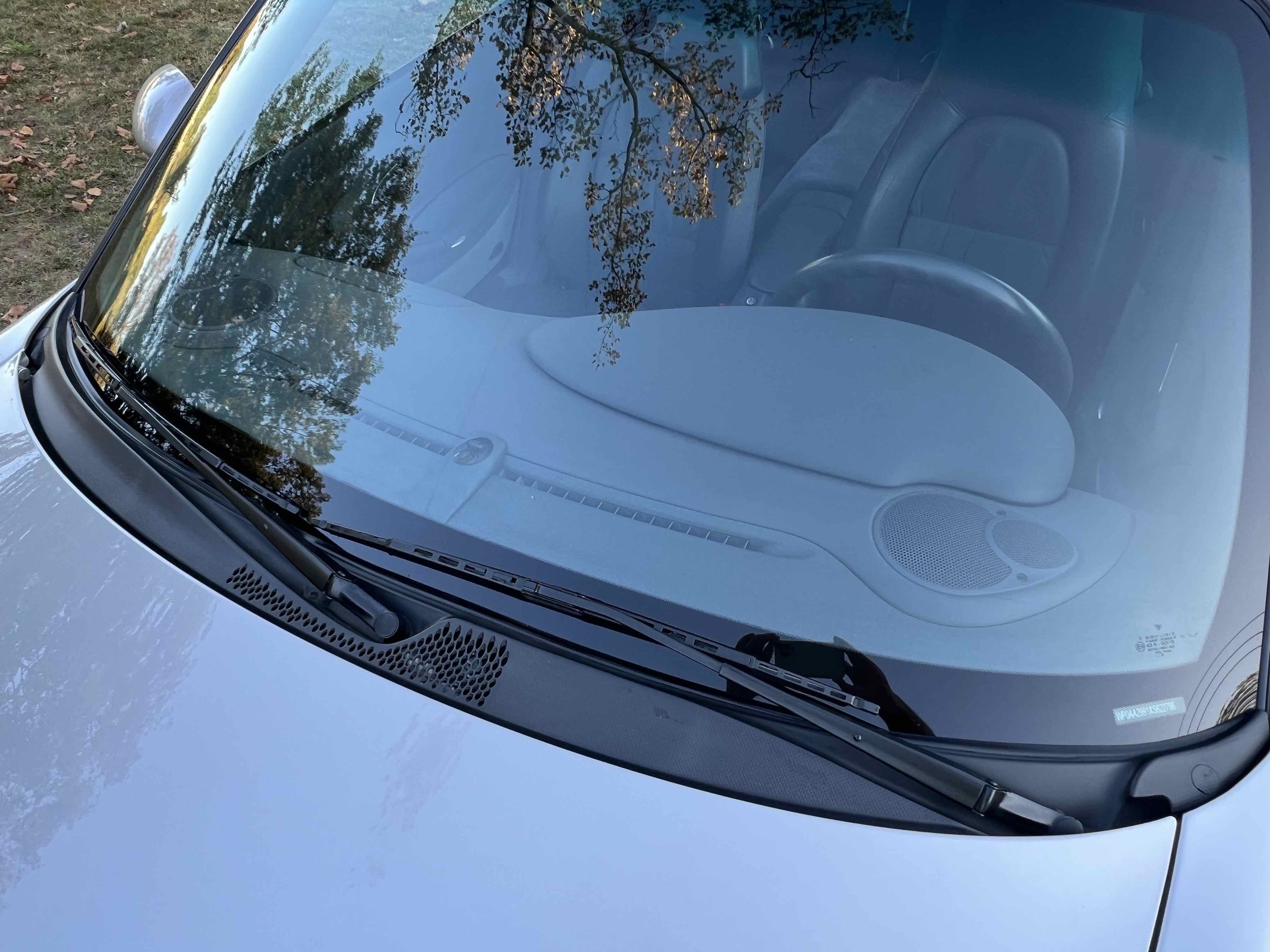 Front windshield of a silver car with reflections of trees. Inside the car, a headrest and a speaker are visible.