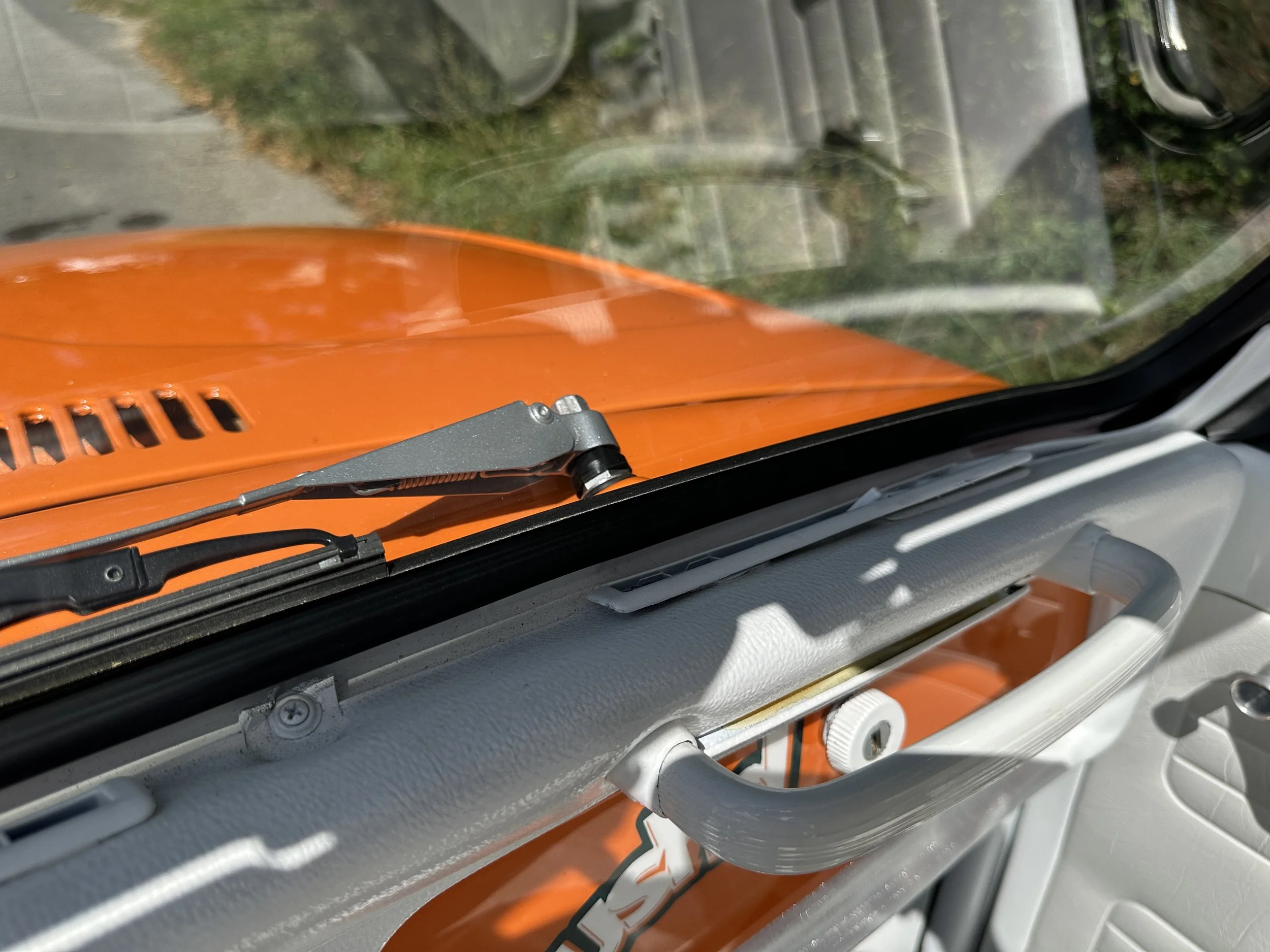 Close-up view of a vehicle dashboard and windshield showing an orange hood with a windshield wiper, and part of the vehicle's door and handle visible inside the vehicle.