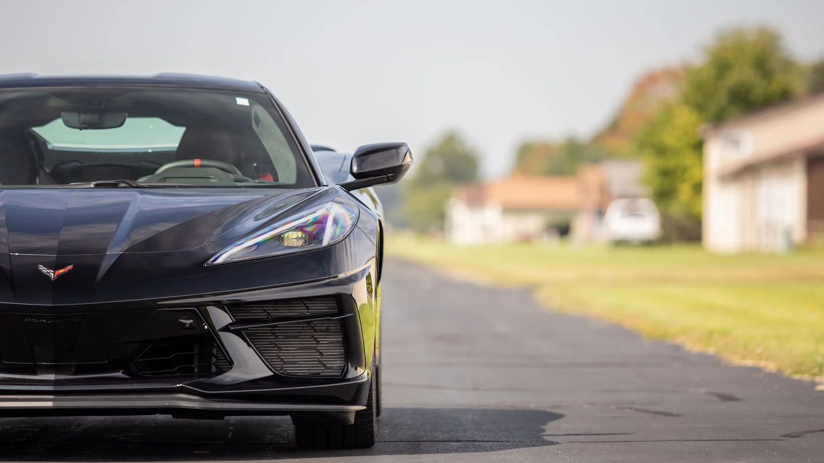 Close-up of the front left side of a black Chevy Corvette sports car parked on a residential street with houses and trees in the background.