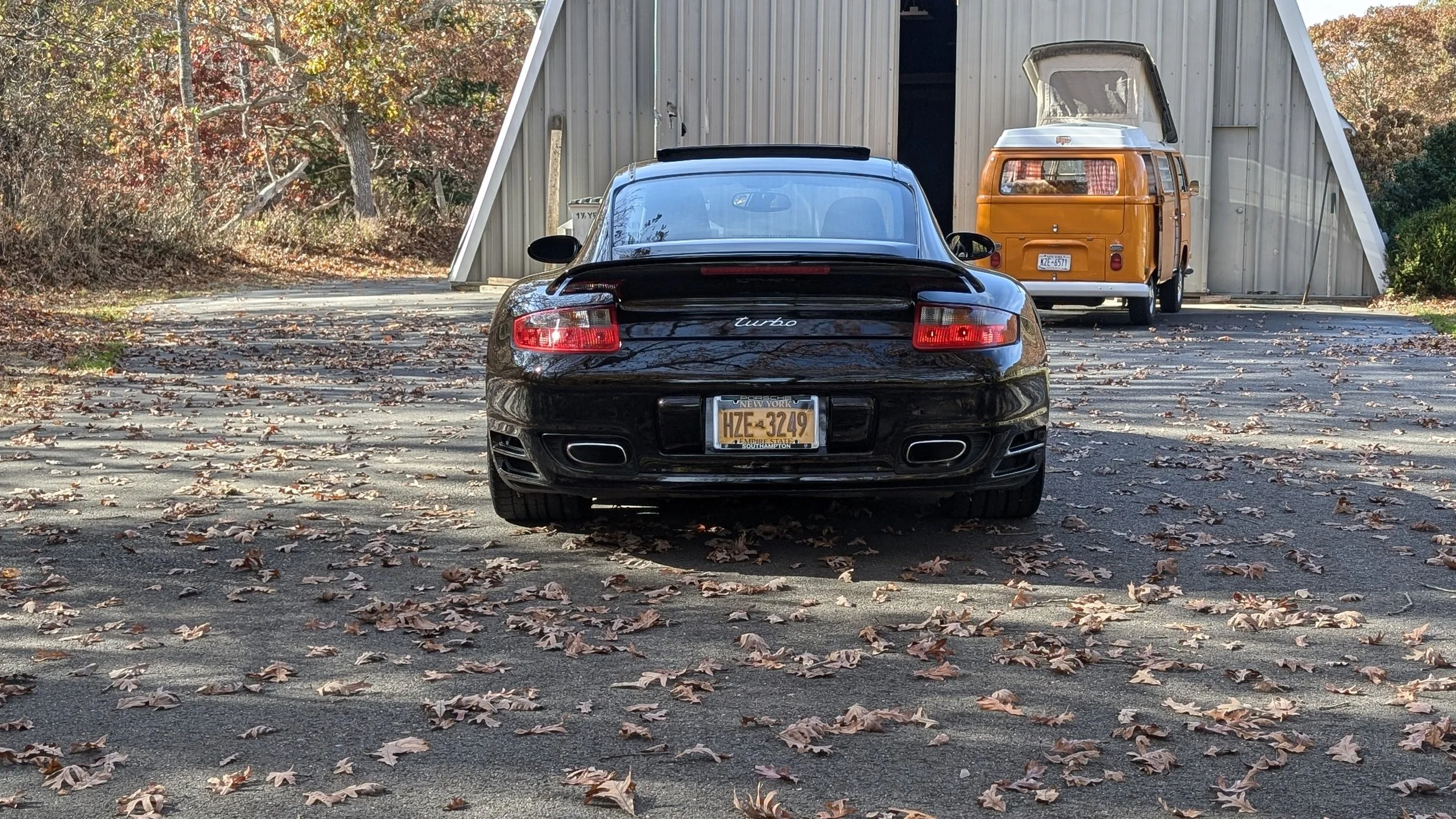 Black Porsche Turbo sports car parked in front of a gray shed with a brown and orange vintage camper trailer in the background, surrounded by fallen autumn leaves.