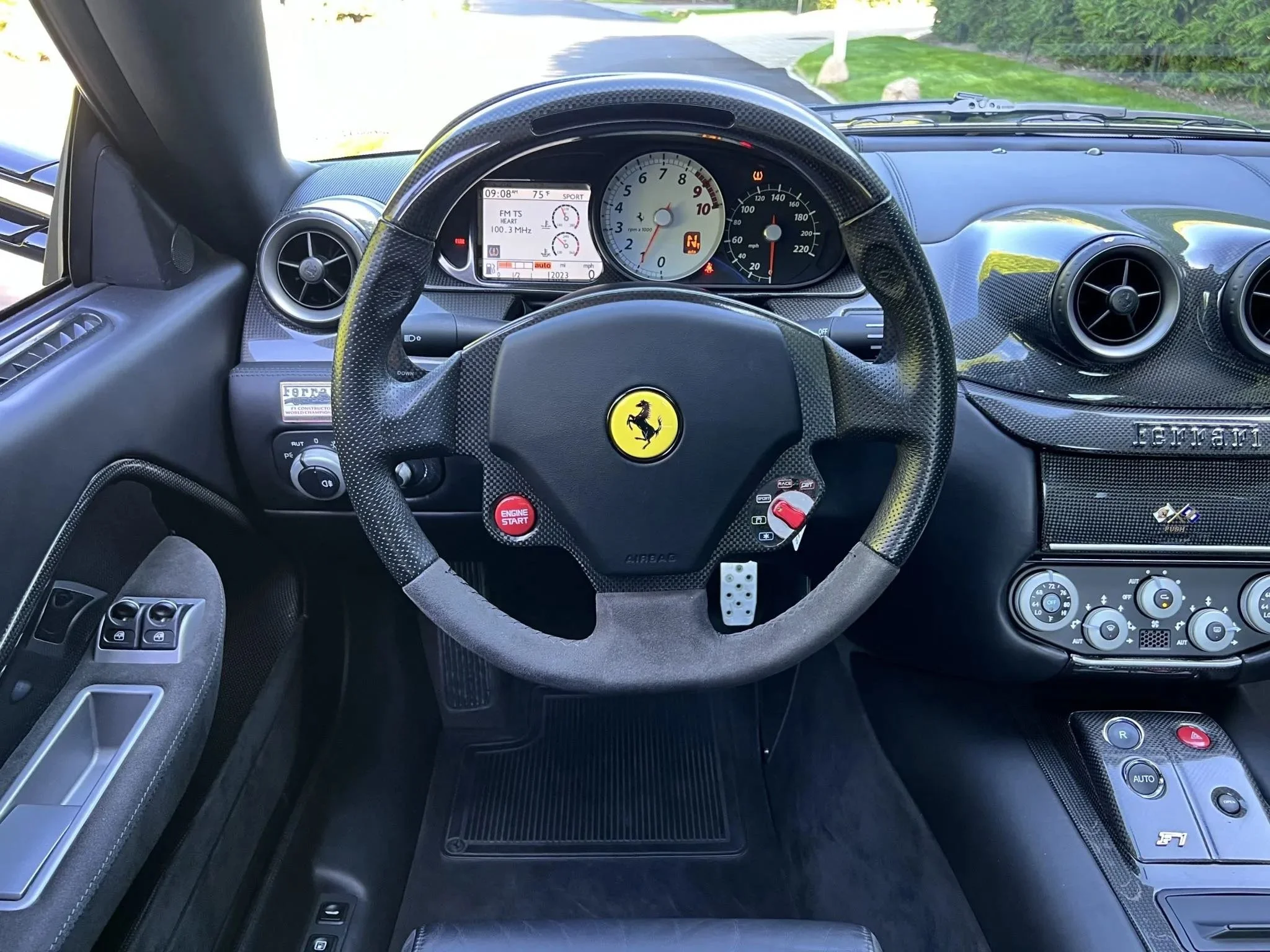 Interior of a Ferrari sports car, showing a steering wheel with Ferrari's prancing horse logo, digital dashboard, climate controls, and carbon fiber accents.