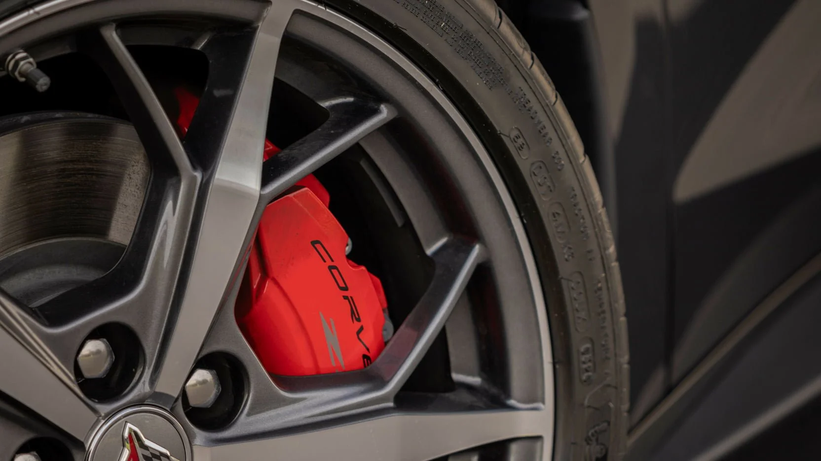 Close-up of a car wheel with a silver alloy rim, black tire, red brake caliper with 'CORVETTE' written on it, and part of a black body panel in the background.