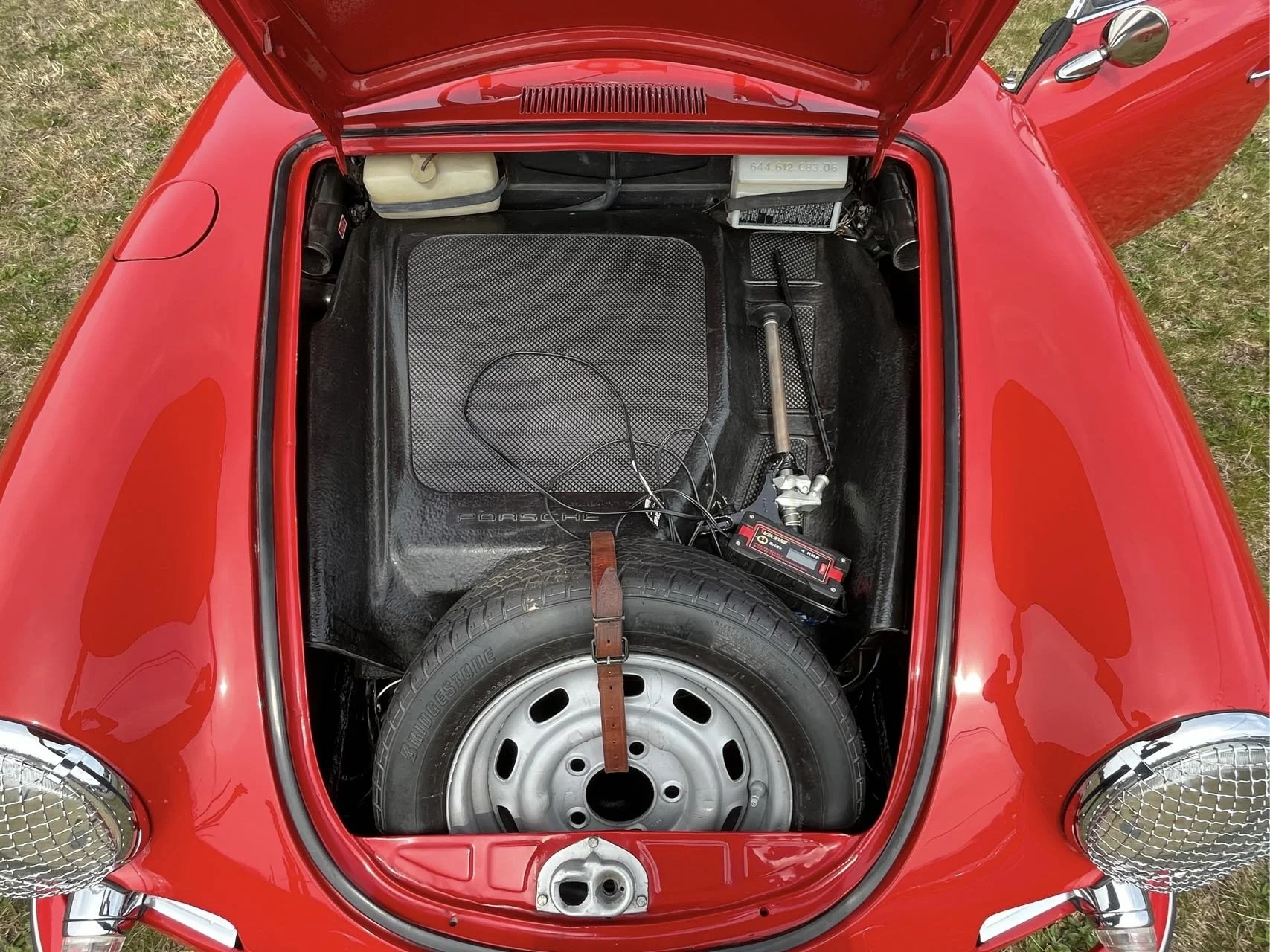 View of the front trunk of a red vintage Porsche sports car, showing a spare tire, a battery pack, and other components.