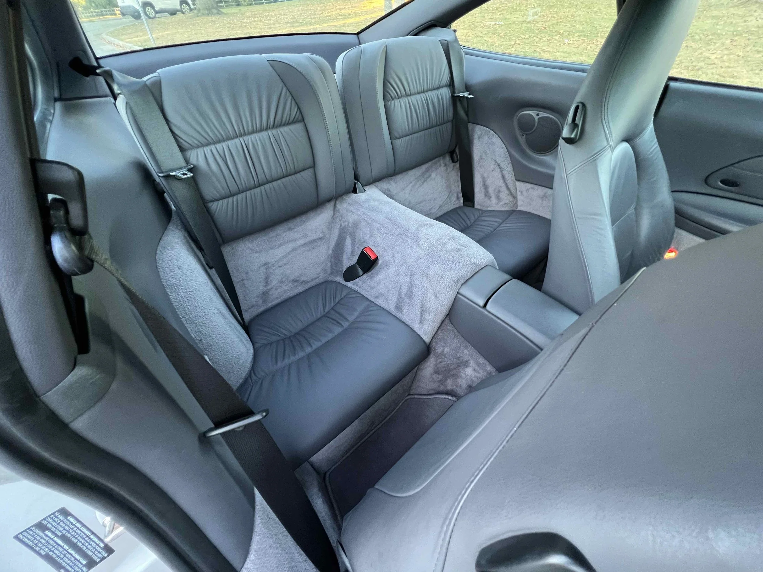 The back seat of a car with gray and black leather seats, seat belts, and a gray carpeted floor. The interior also features side panels with speakers and windows showing an outdoor grassy area.