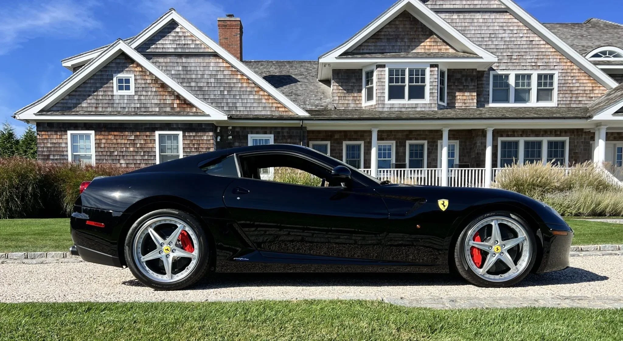 Black Ferrari sports car parked on a gravel driveway in front of a large house with a shingle roof, white porch, and multiple windows, under a clear blue sky.