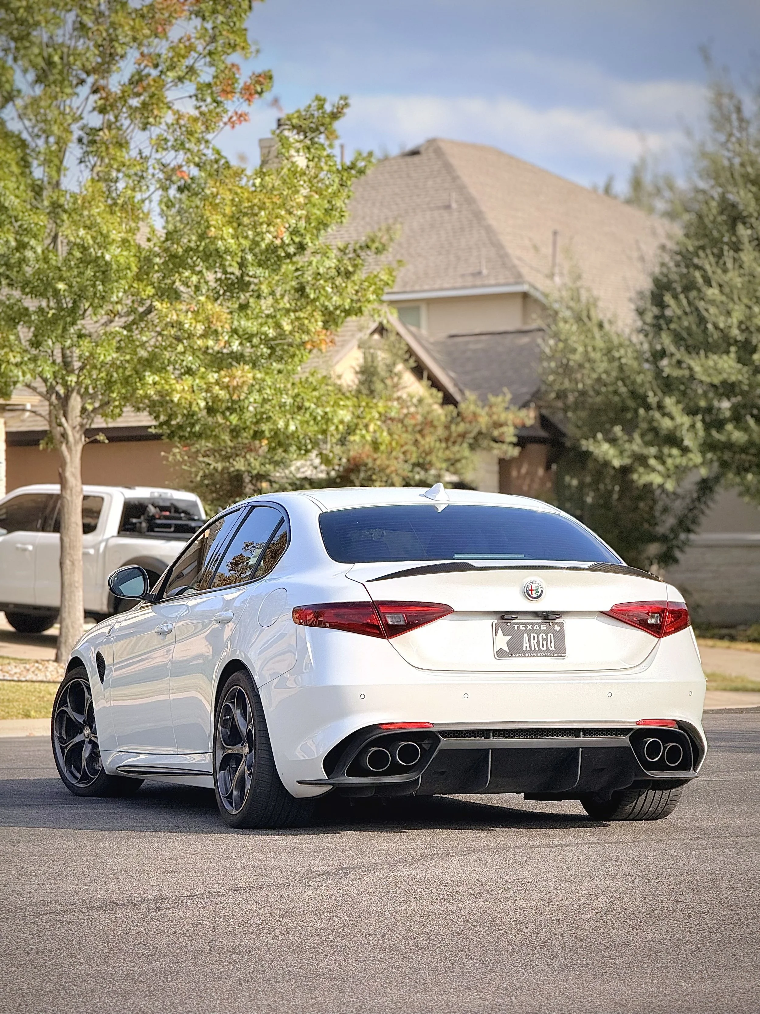 A white Alfa Romeo Giulia parked on a suburban street with houses, trees, clear sky, and another vehicle in background.