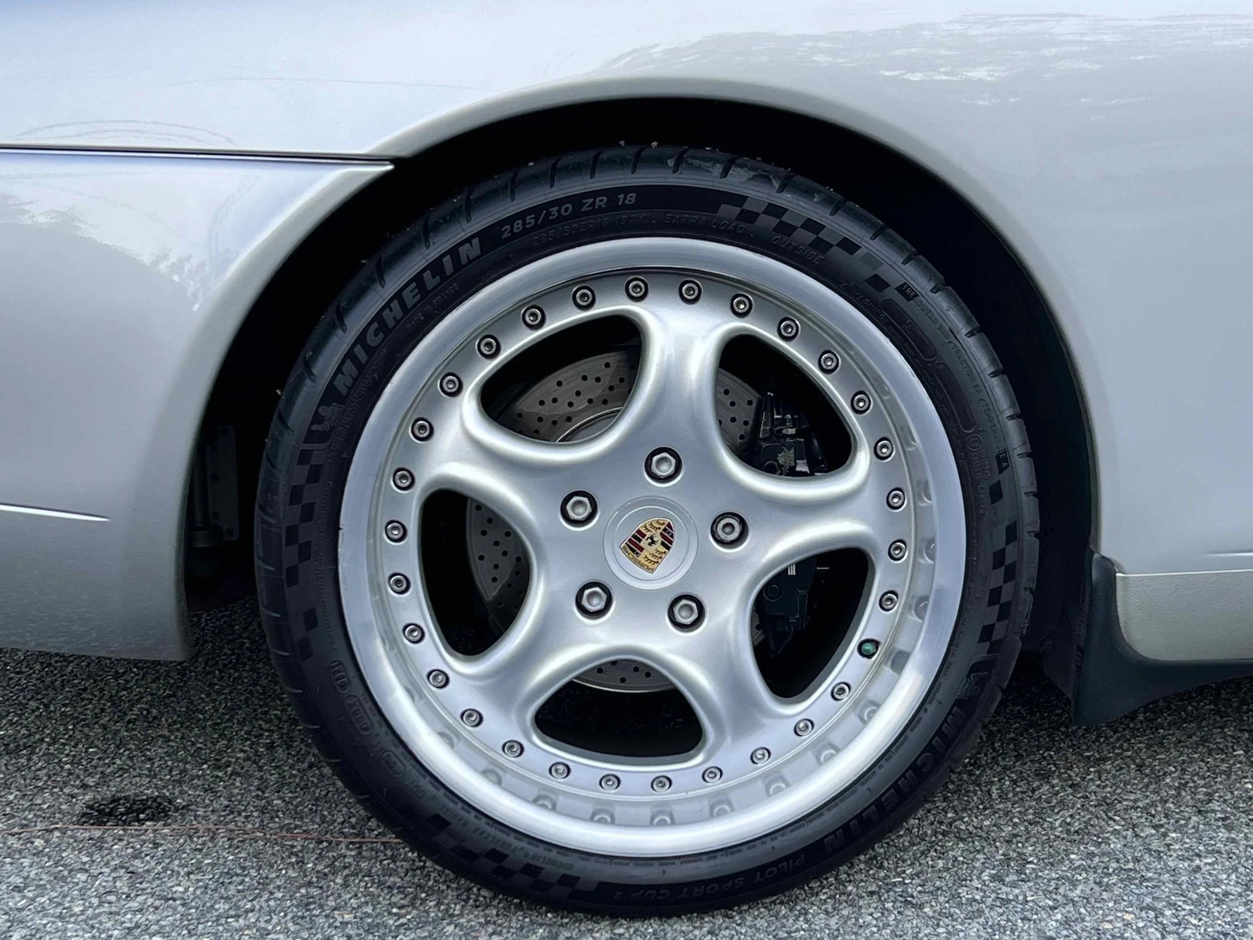 Close-up of a silver Porsche car wheel with Michelin tires and a black and gold Porsche emblem in the center.
