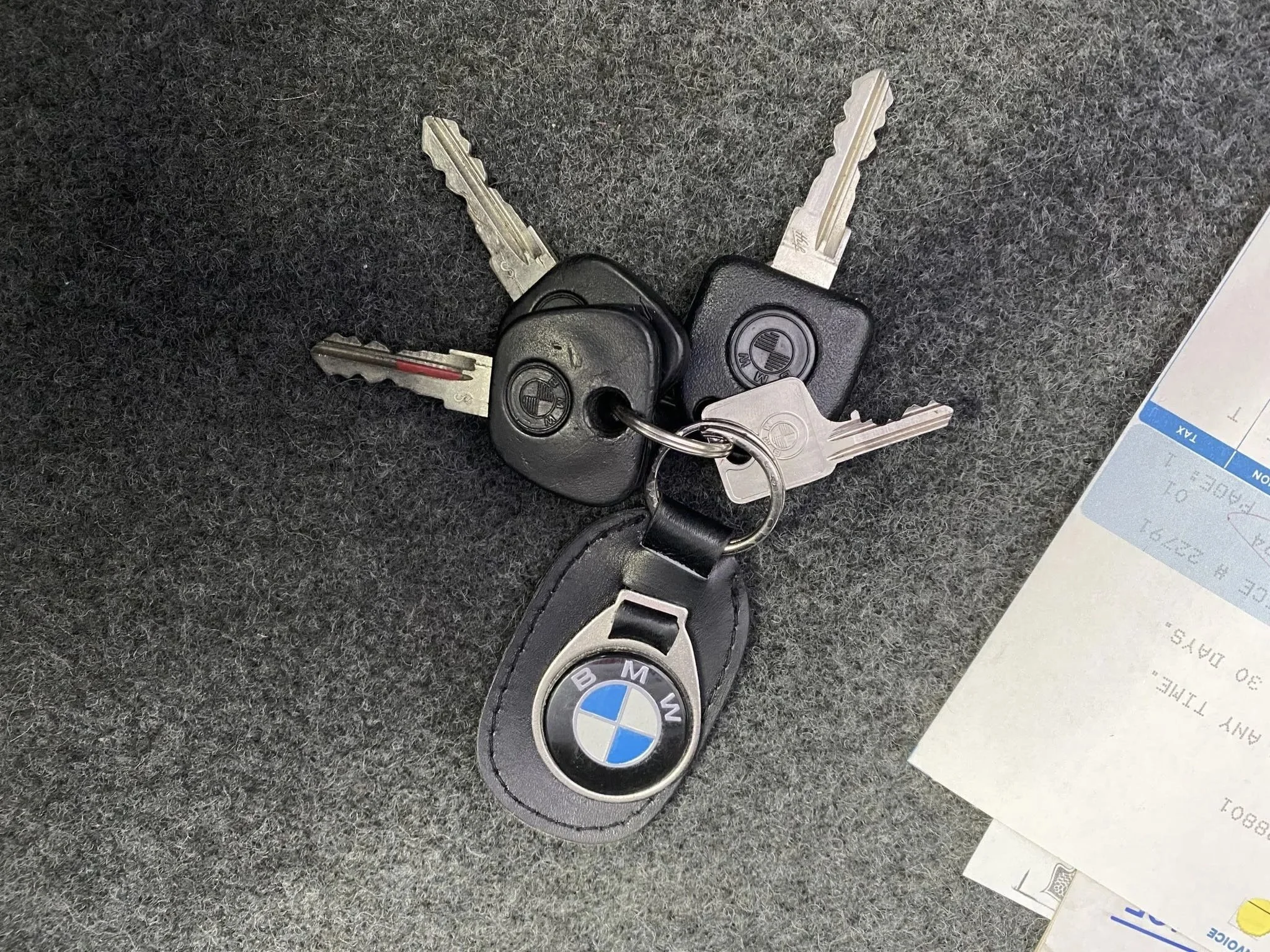 A set of keys on a BMW keychain, including BMW key fobs, placed on a dark textured surface with part of a printed paper visible nearby.
