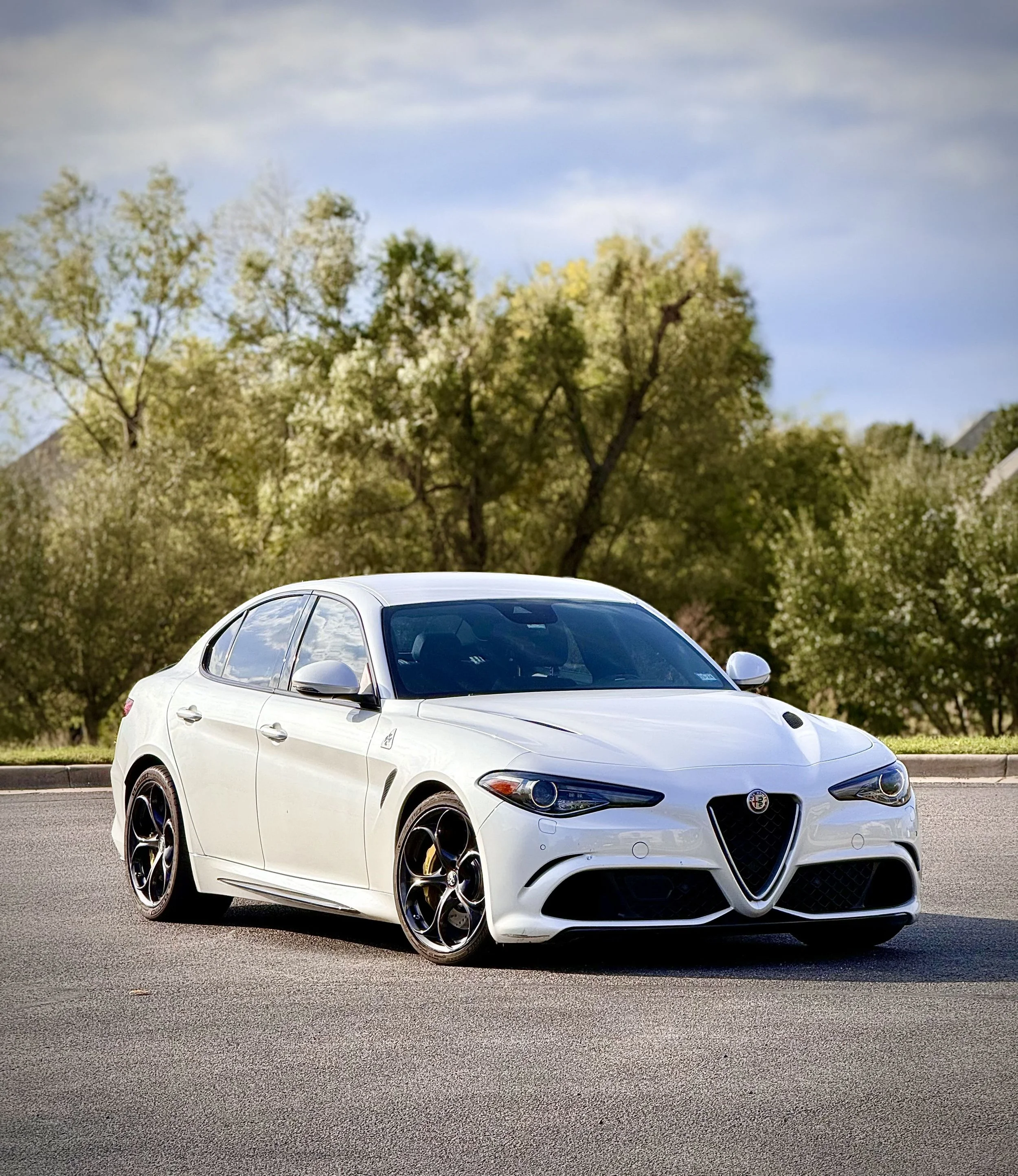 White sports car with black wheels on a paved road, trees and a partly cloudy sky in the background.