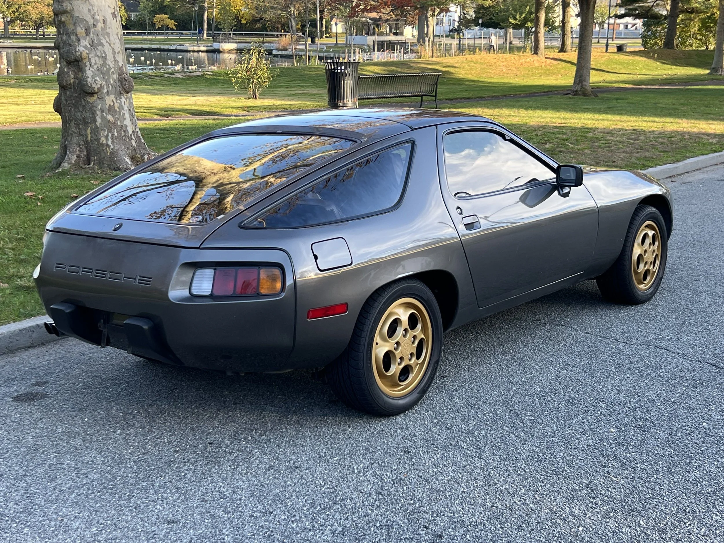 A classic gray Porsche sports car with gold wheels parked on a street near a park with trees, grass, a bench, and a pond.