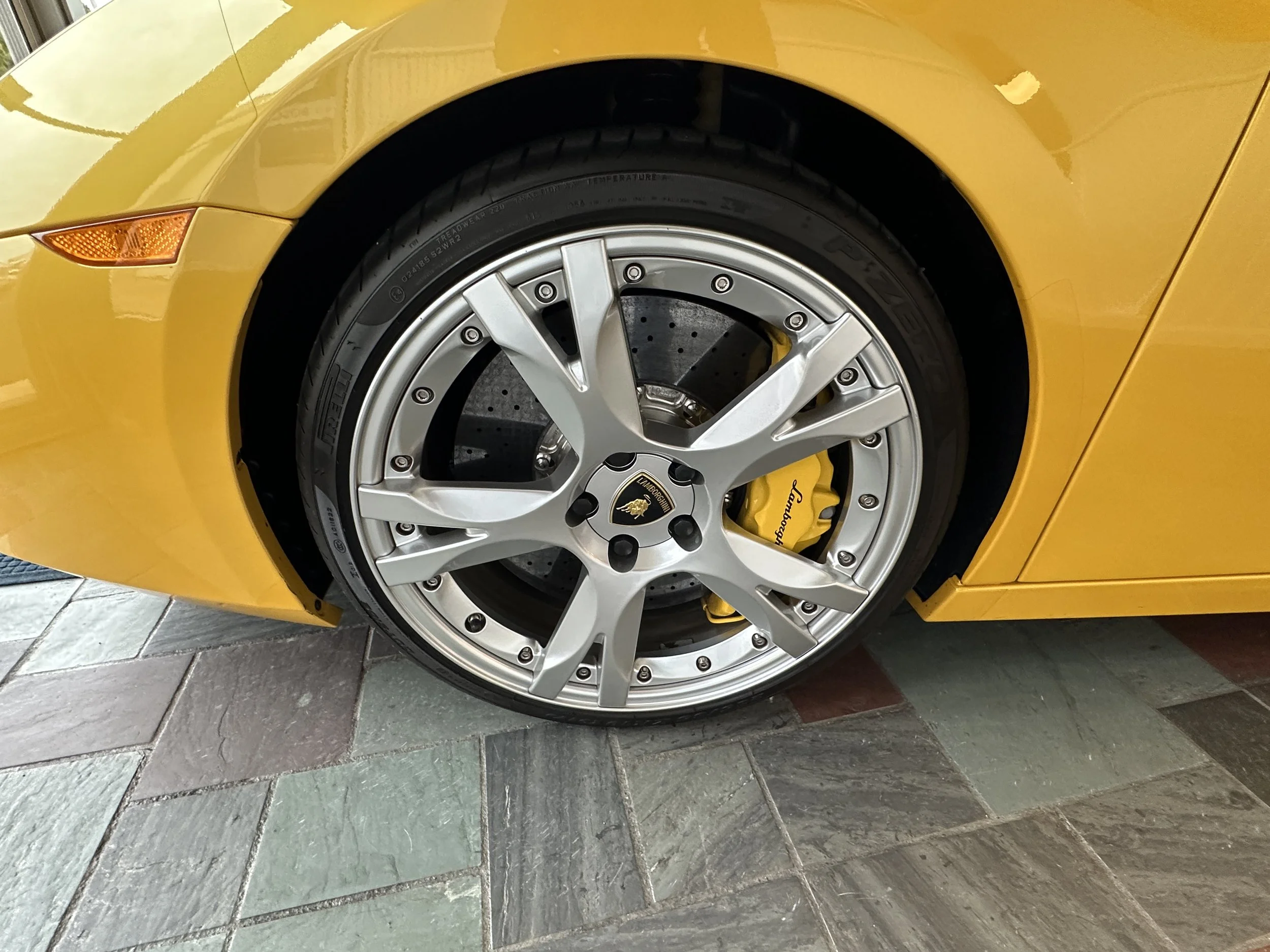 Close-up of a yellow Lamborghini car wheel showing a silver rim, yellow brake caliper with Lamborghini logo, and treaded tire, on tiled floor.