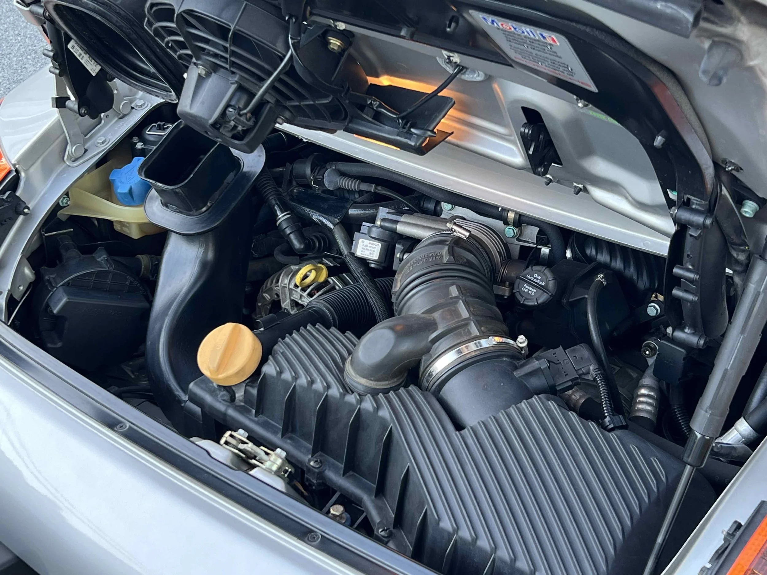 Car engine compartment with various engine components, hoses, and a black plastic cover.