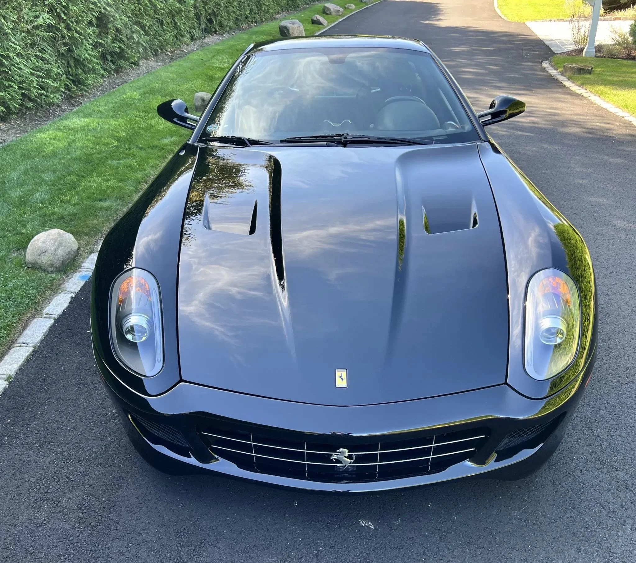 Black Ferrari sports car with hood vents, parked on asphalt road next to a grassy area with rocks and green bushes, shot from the front angle.