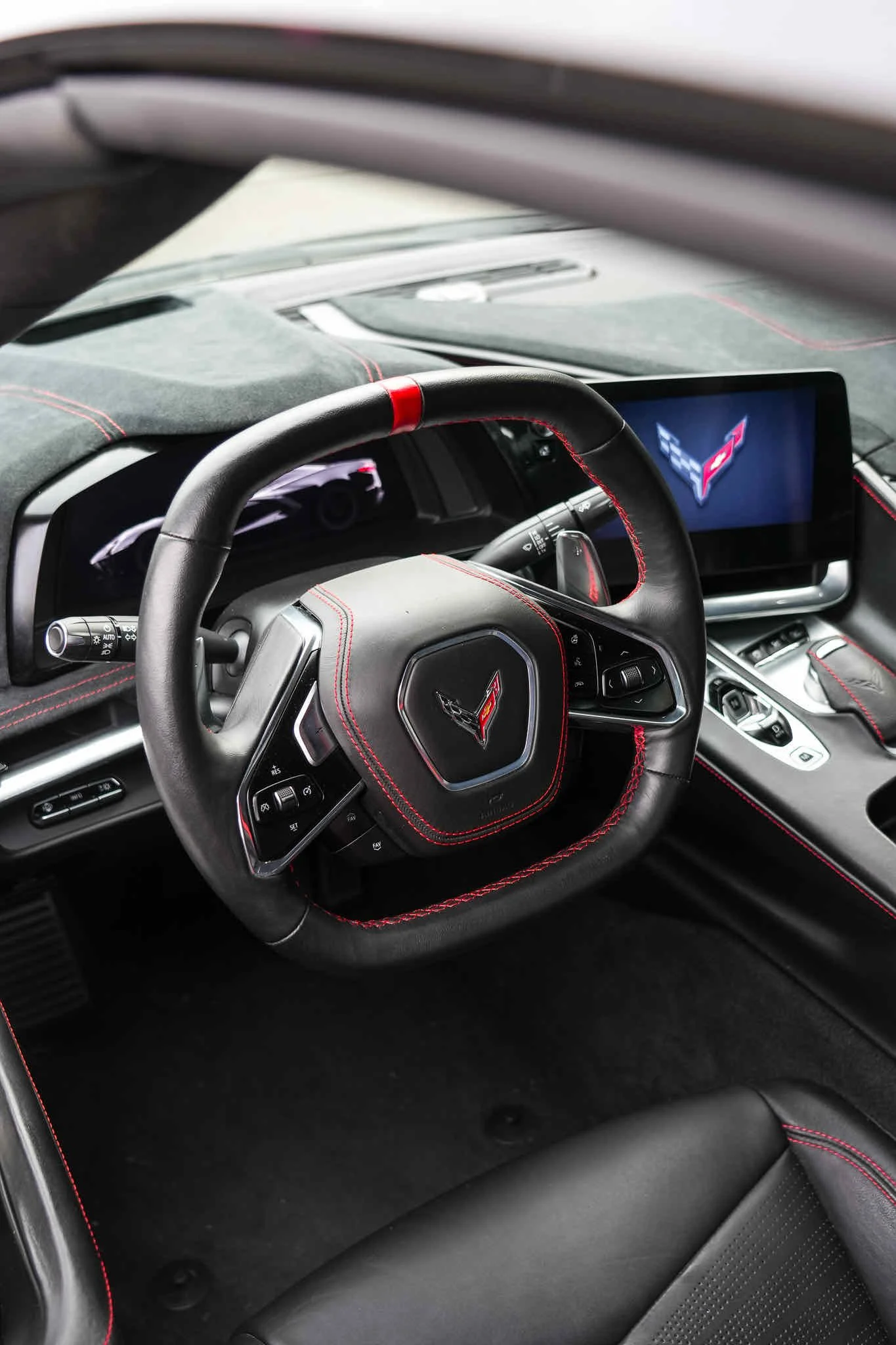 Close-up of the interior of a modern sports car, showing a black leather steering wheel with red stitching, a digital dashboard, and an infotainment screen displaying a Chevrolet Corvette logo.