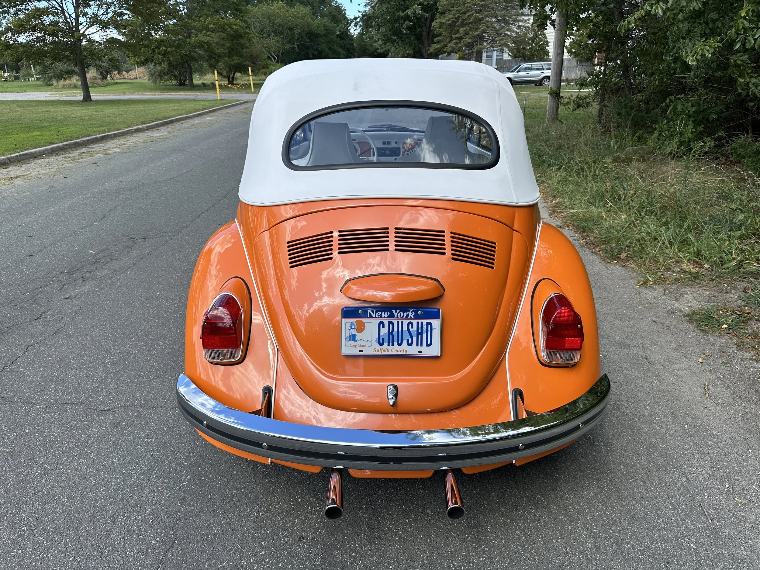 Rear view of a vintage orange Volkswagen Beetle with a white soft top convertible roof parked on a roadside, displaying a New York license plate reading 'CRUSHD'.