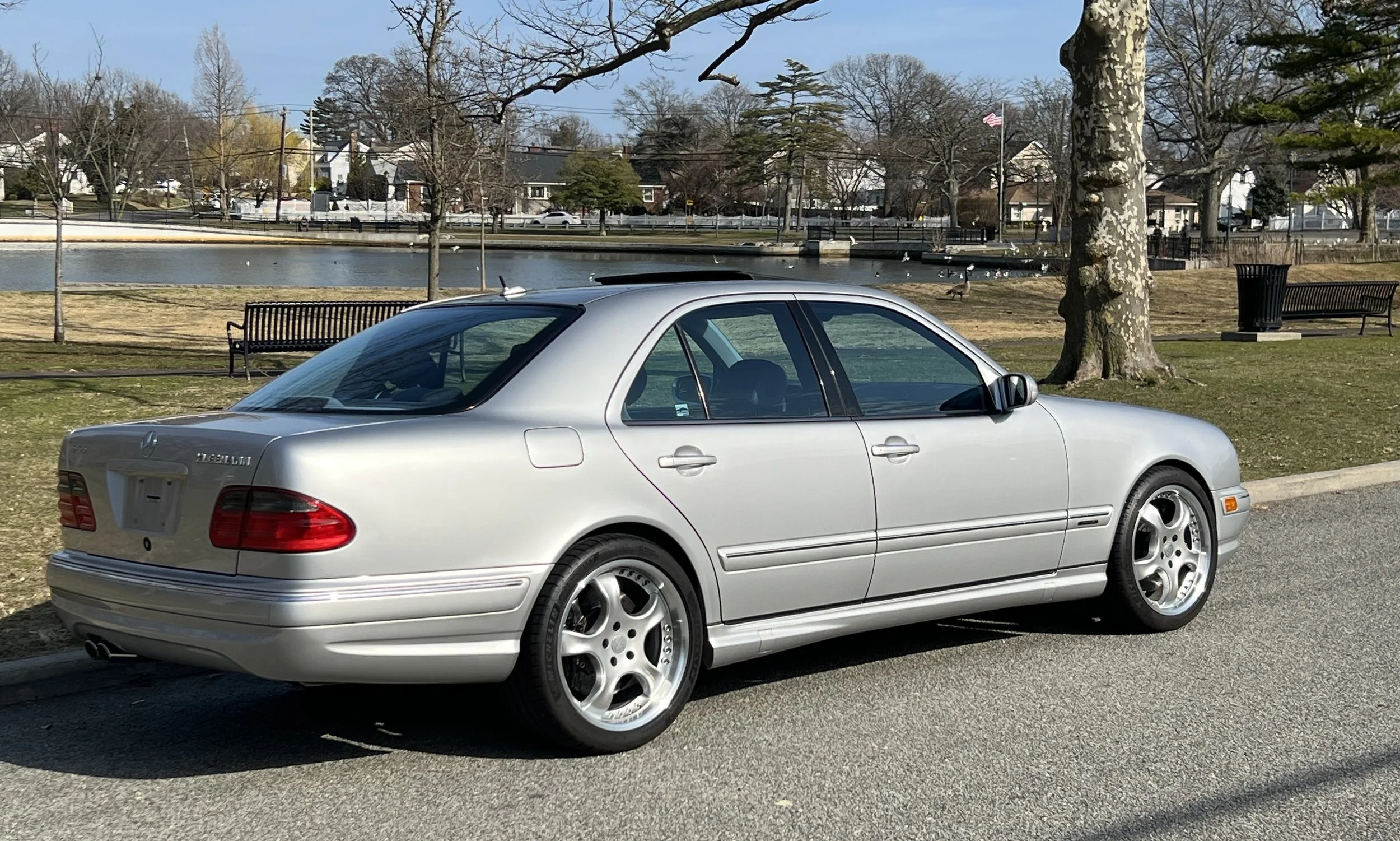 A silver Mercedes-Benz coupe parked near a park with trees, benches, and a pond in the background.
