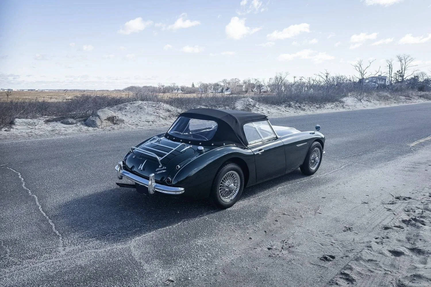 A vintage black convertible sports car parked on the side of a rural road with a sandy, shrub-covered landscape in the background and a partly cloudy sky.