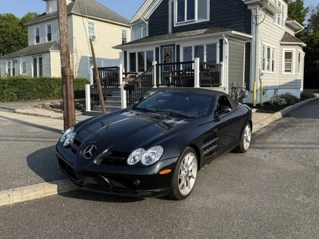 Black Mercedes-Benz convertible parked on the street in front of a house with a porch, steps, and multiple windows.