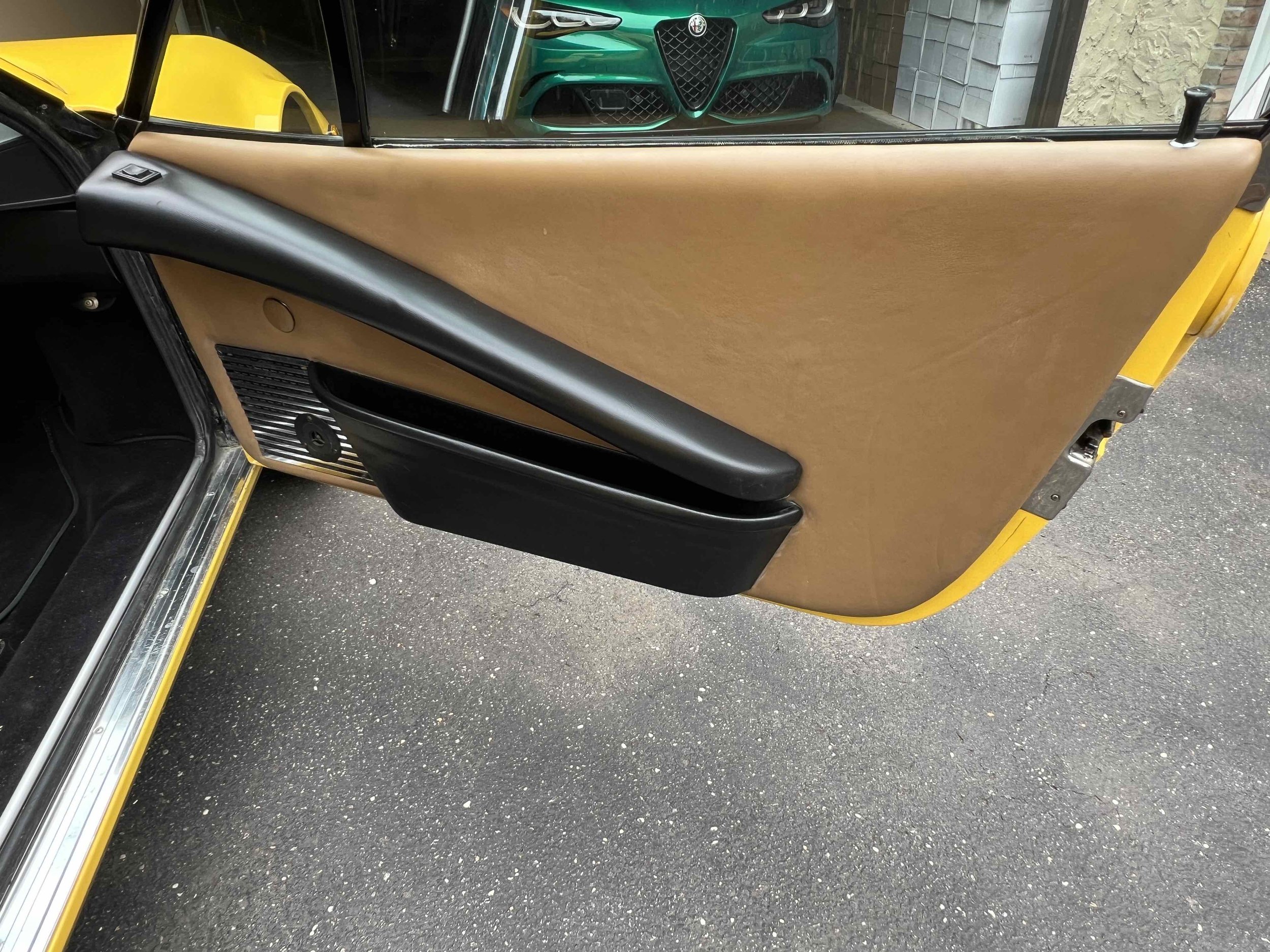 Interior car door with tan upholstery, black armrest, and door handle; part of yellow exterior visible outside.