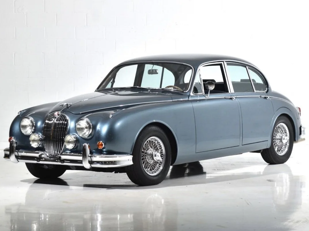 A classic vintage blue car with chrome details and wire-spoke wheels parked indoors against a plain white background.