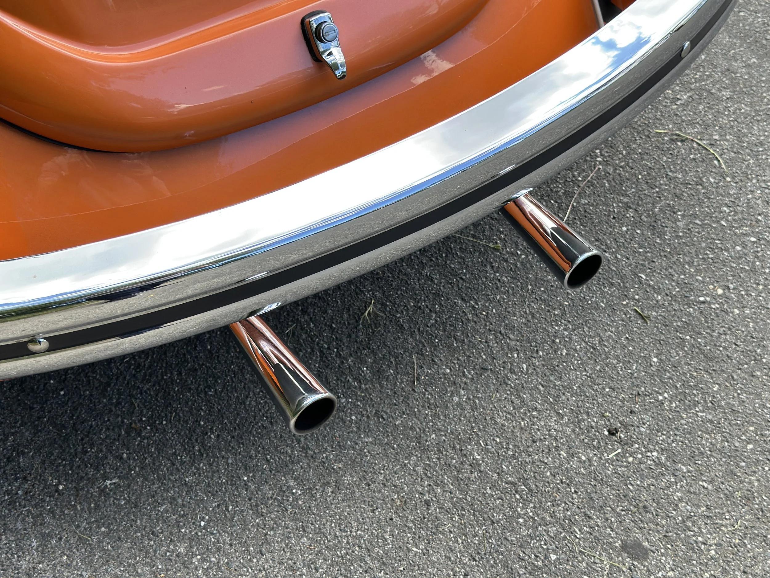 Close-up of the rear end of a vintage orange car showing dual exhaust pipes, a chrome bumper, and part of the trunk lid.