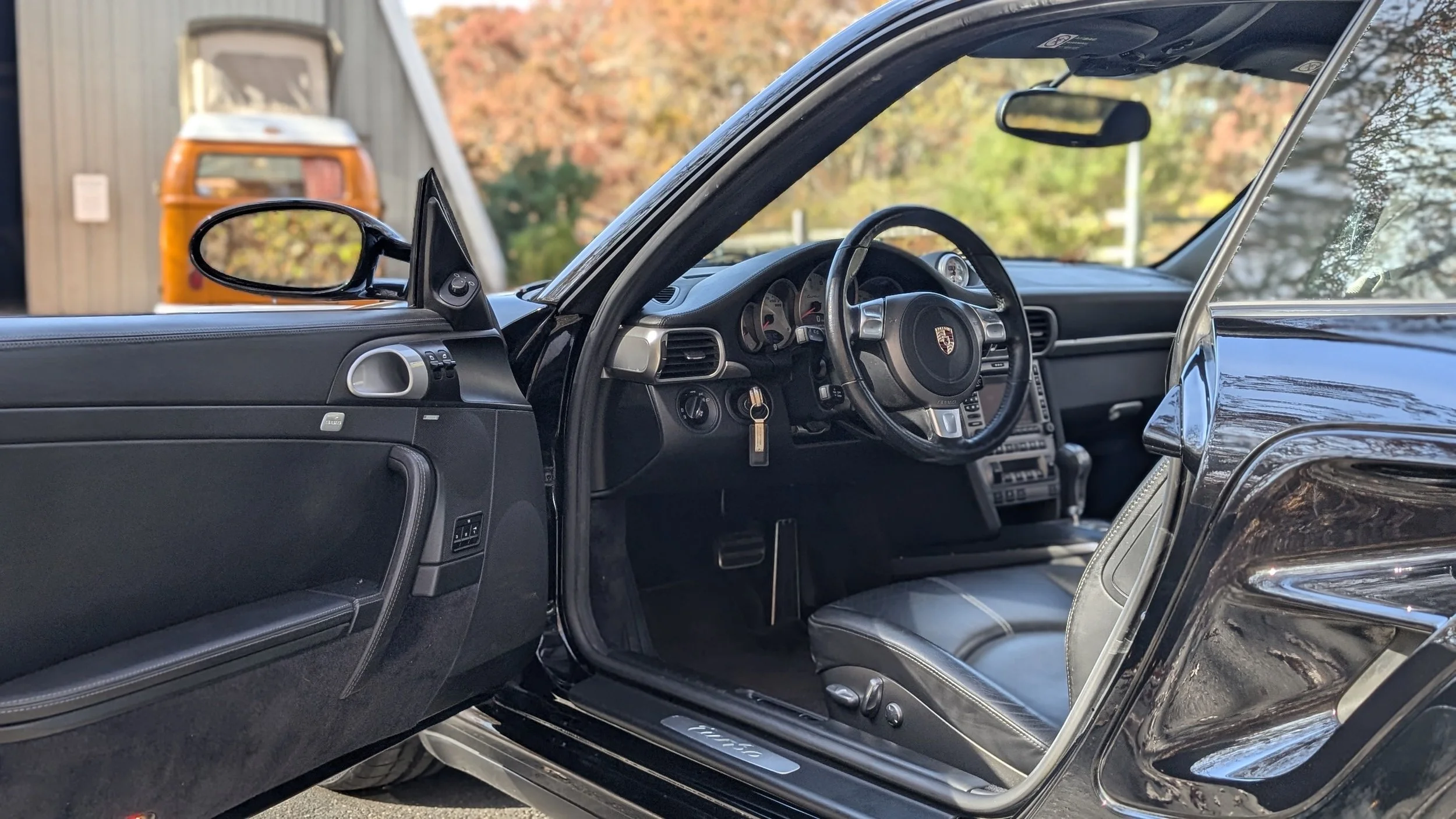 Inside view of a black Porsche car with the driver's side door open, showing the steering wheel, dashboard, and front seat, with an orange vintage van in the background outside.