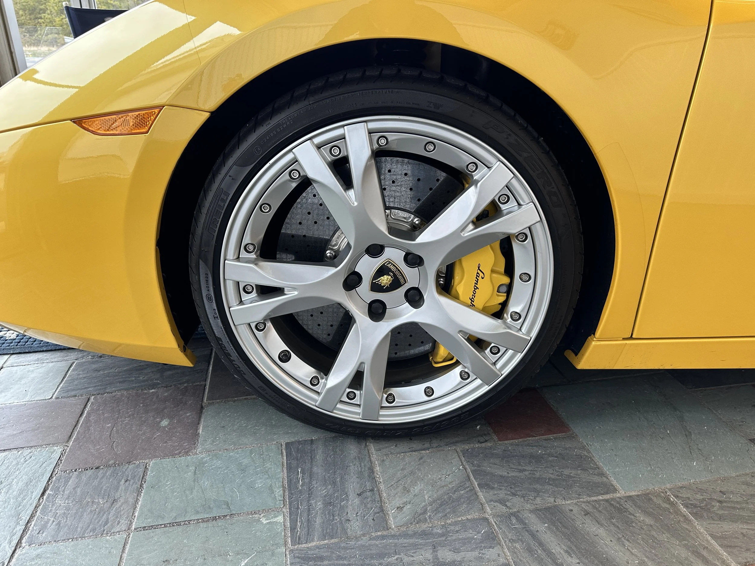 Close-up of a yellow Lamborghini sports car's front wheel, showing a multi-spoke alloy rim, a large brake disc, and a yellow brake caliper with Lamborghini branding.