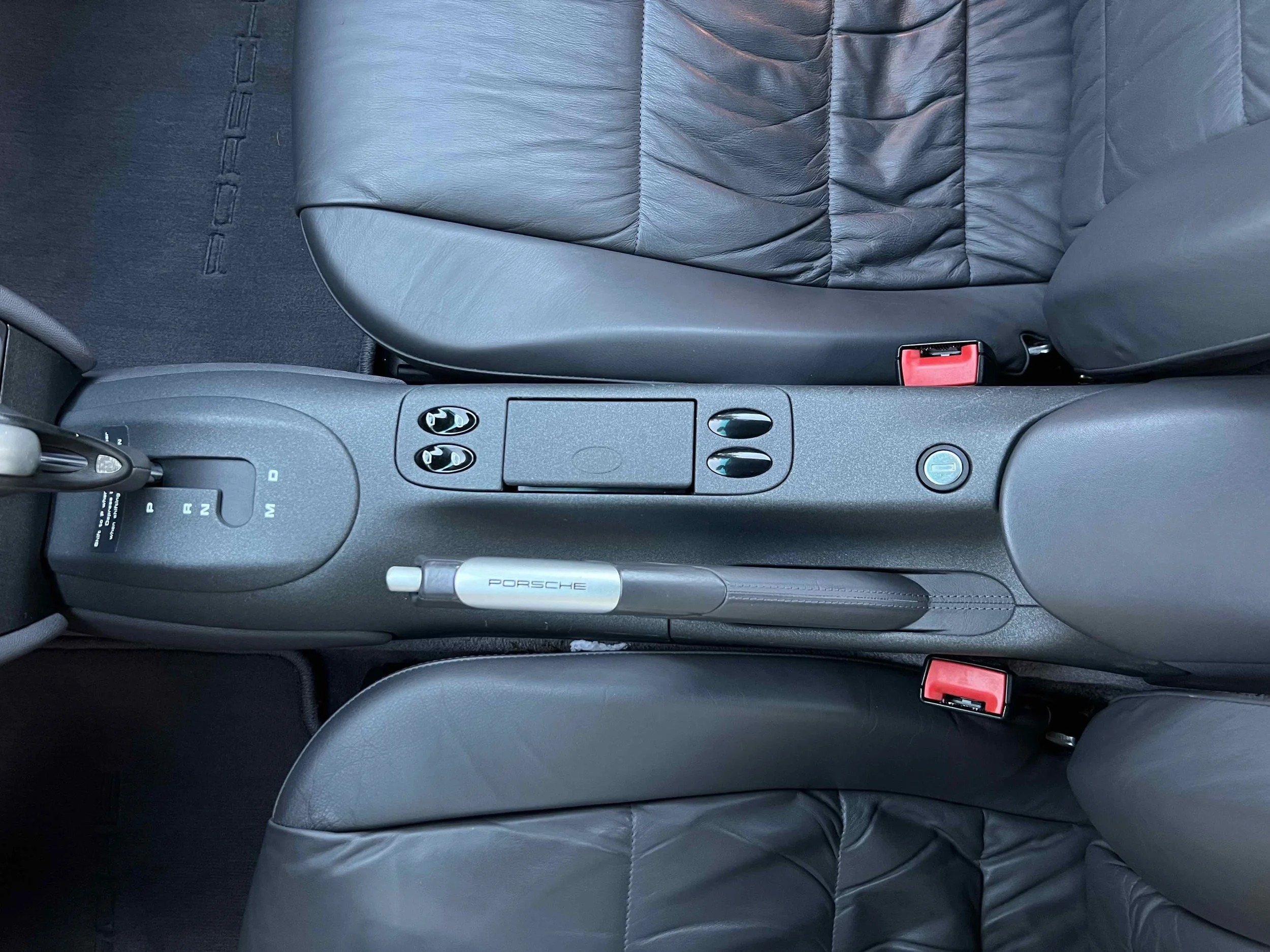 Top-down view of the center console and front seats of a black Porsche car, showing gear shift, control buttons, and seat belts.
