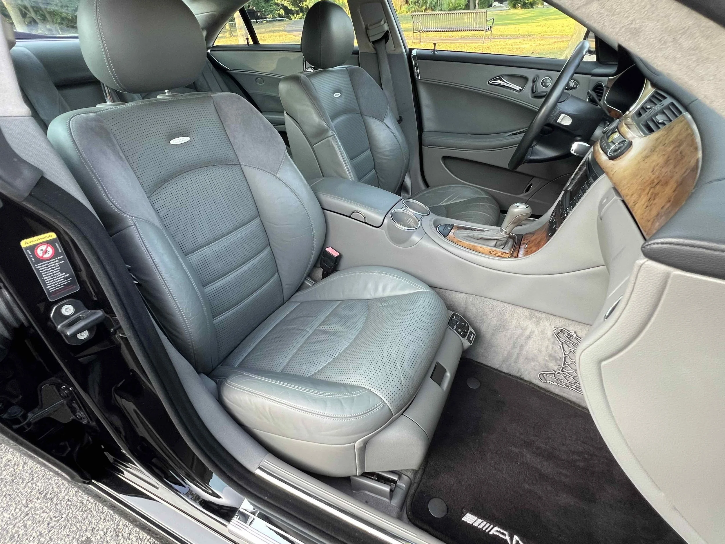 The interior of a car showing gray leather seats, a wood-paneled dashboard, and center console with cupholders, with a parking lot and grassy area visible through the windows.