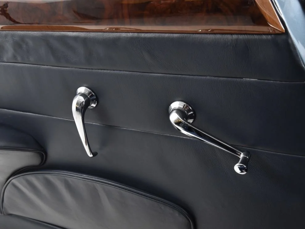 Close-up of a car door interior featuring black leather upholstery and two chrome window cranks.
