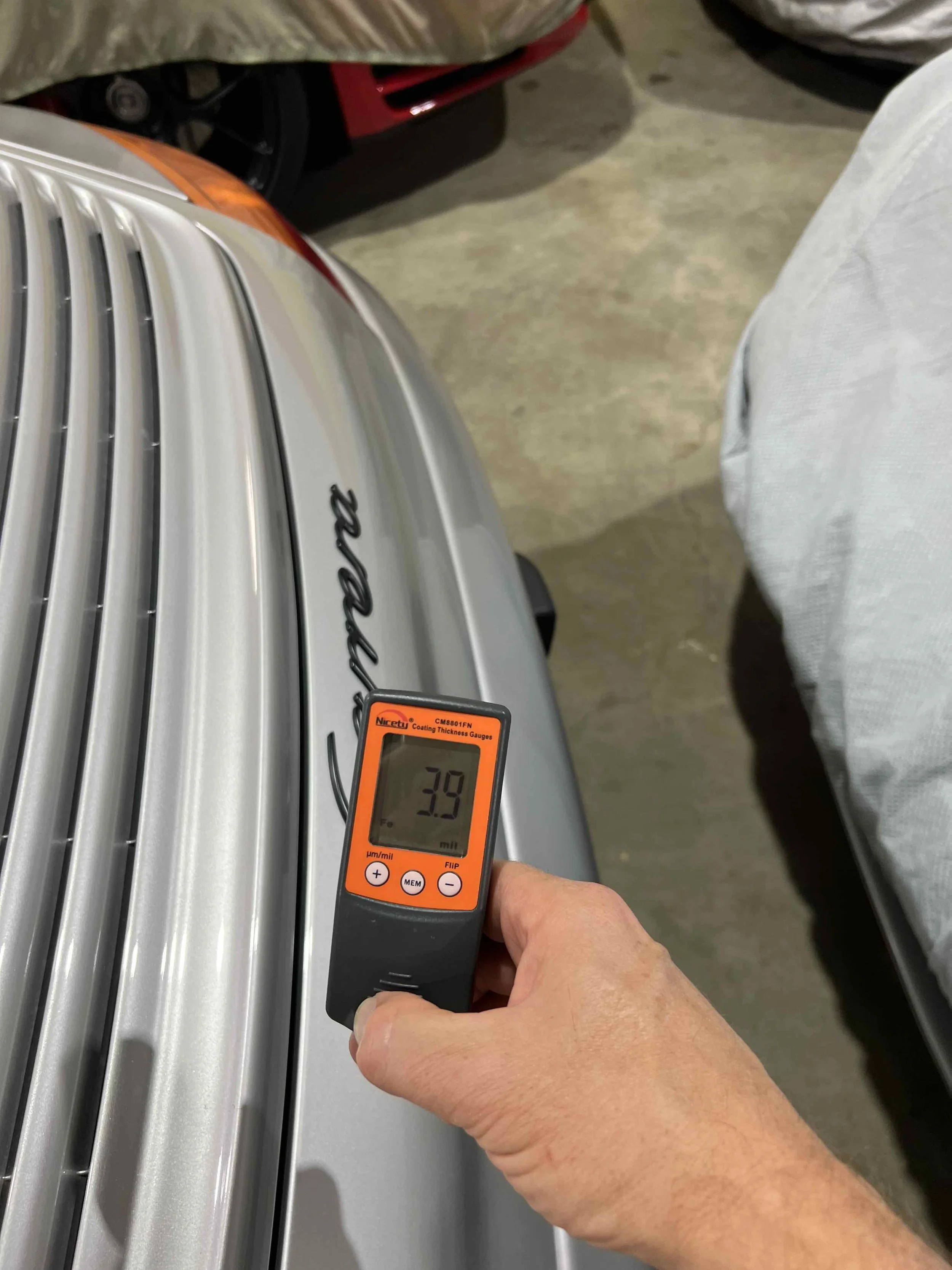 Person measuring paint thickness on a silver car's hood using an orange digital coating thickness gauge.