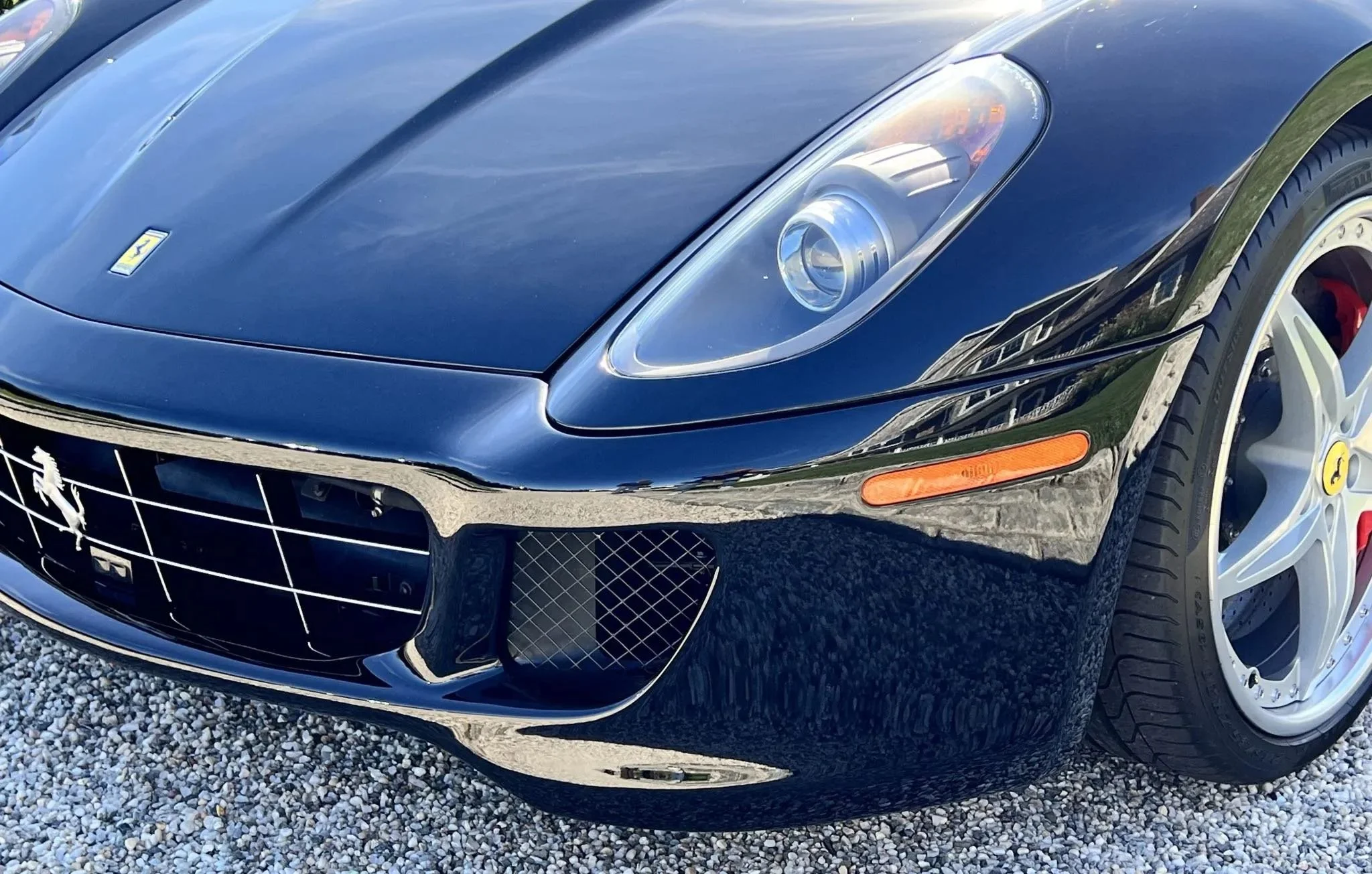 Front partial view of a black Ferrari sports car, showing the headlight, front grille with the prancing horse emblem, and a portion of the tire and wheel.