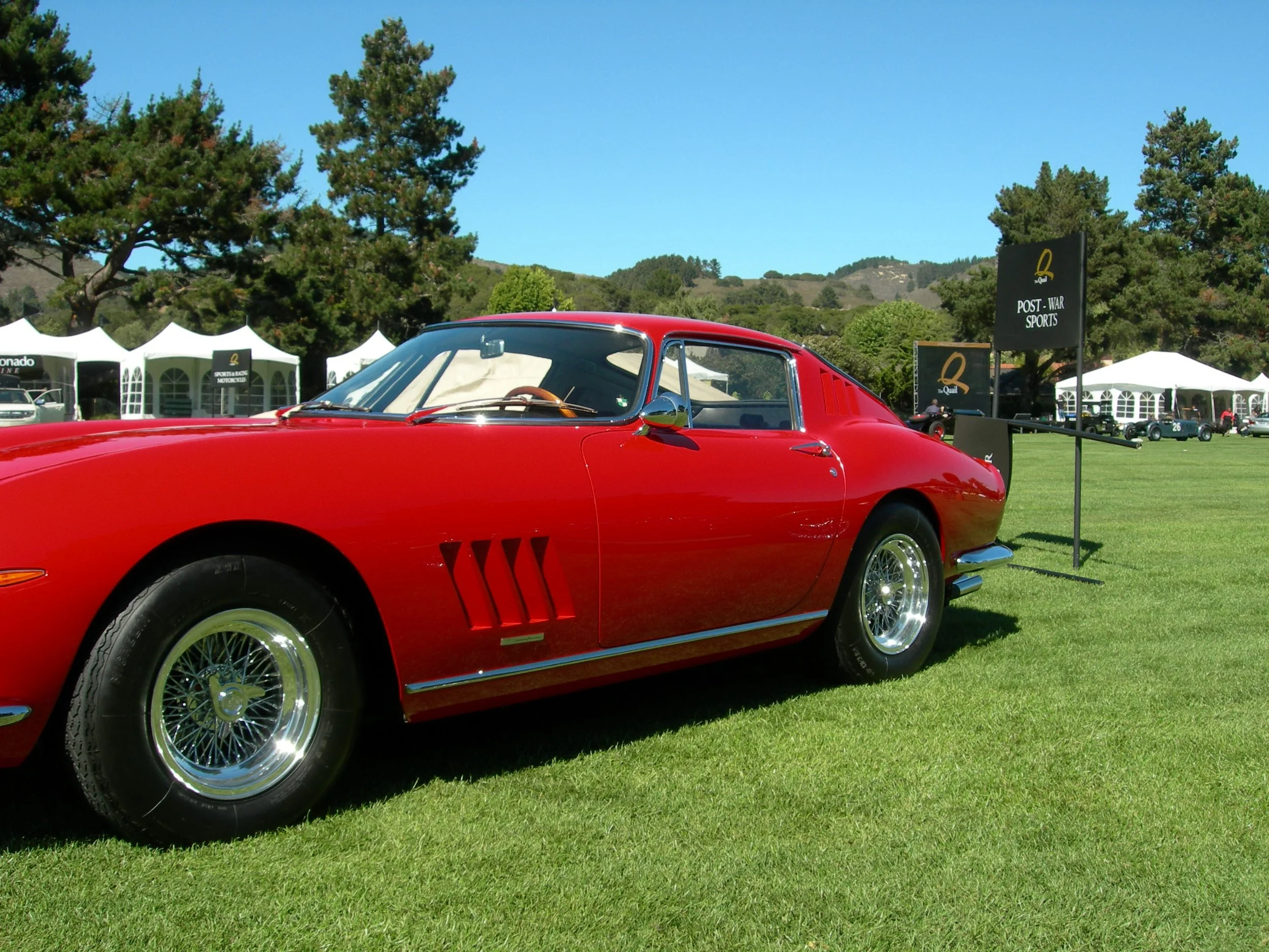 A red vintage sports car displayed at an outdoor automobile show on a grassy field, with tents and trees in the background.