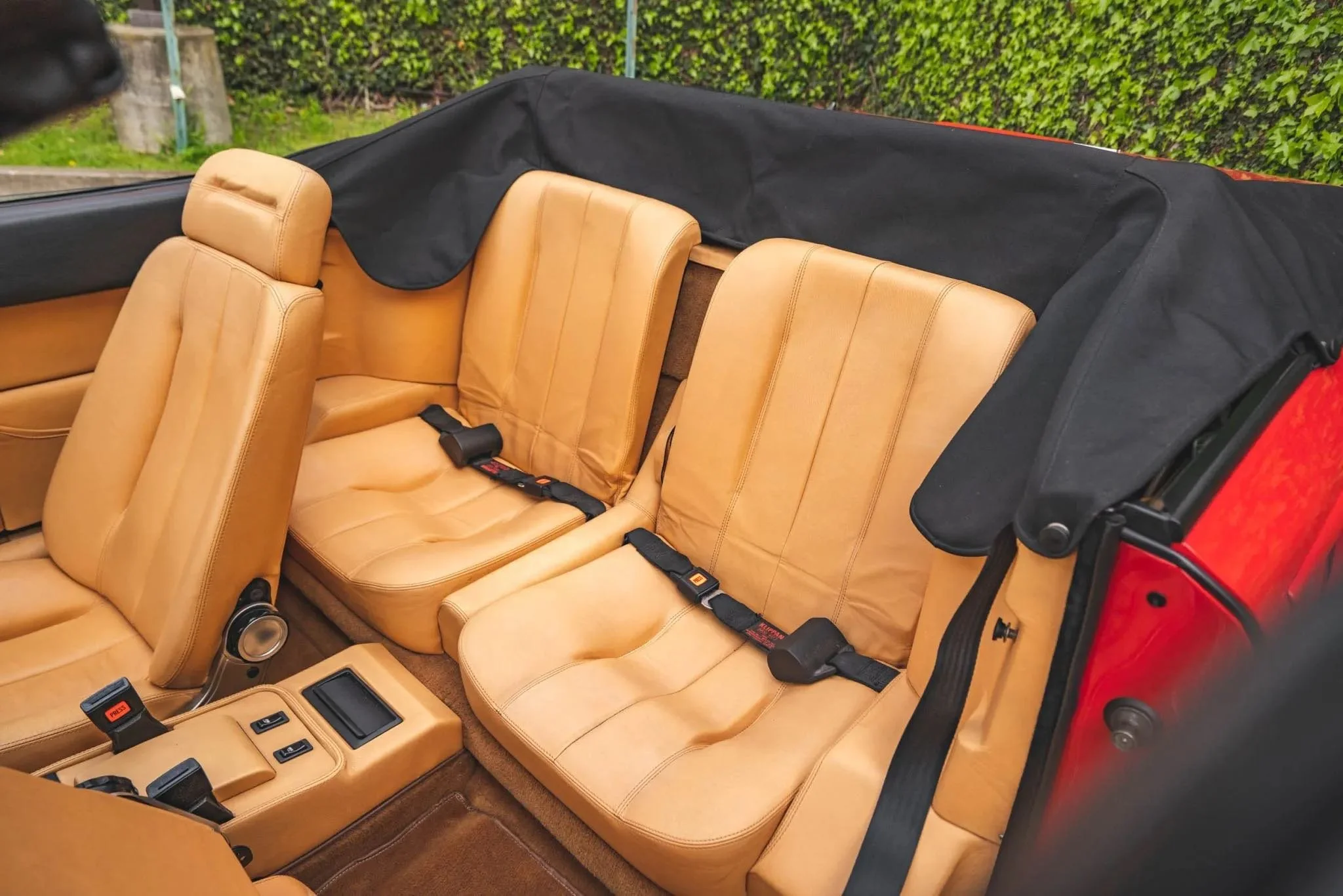Interior of a car showing tan leather seats and seat belts, with a black soft top partially folded back, outside greenery visible.