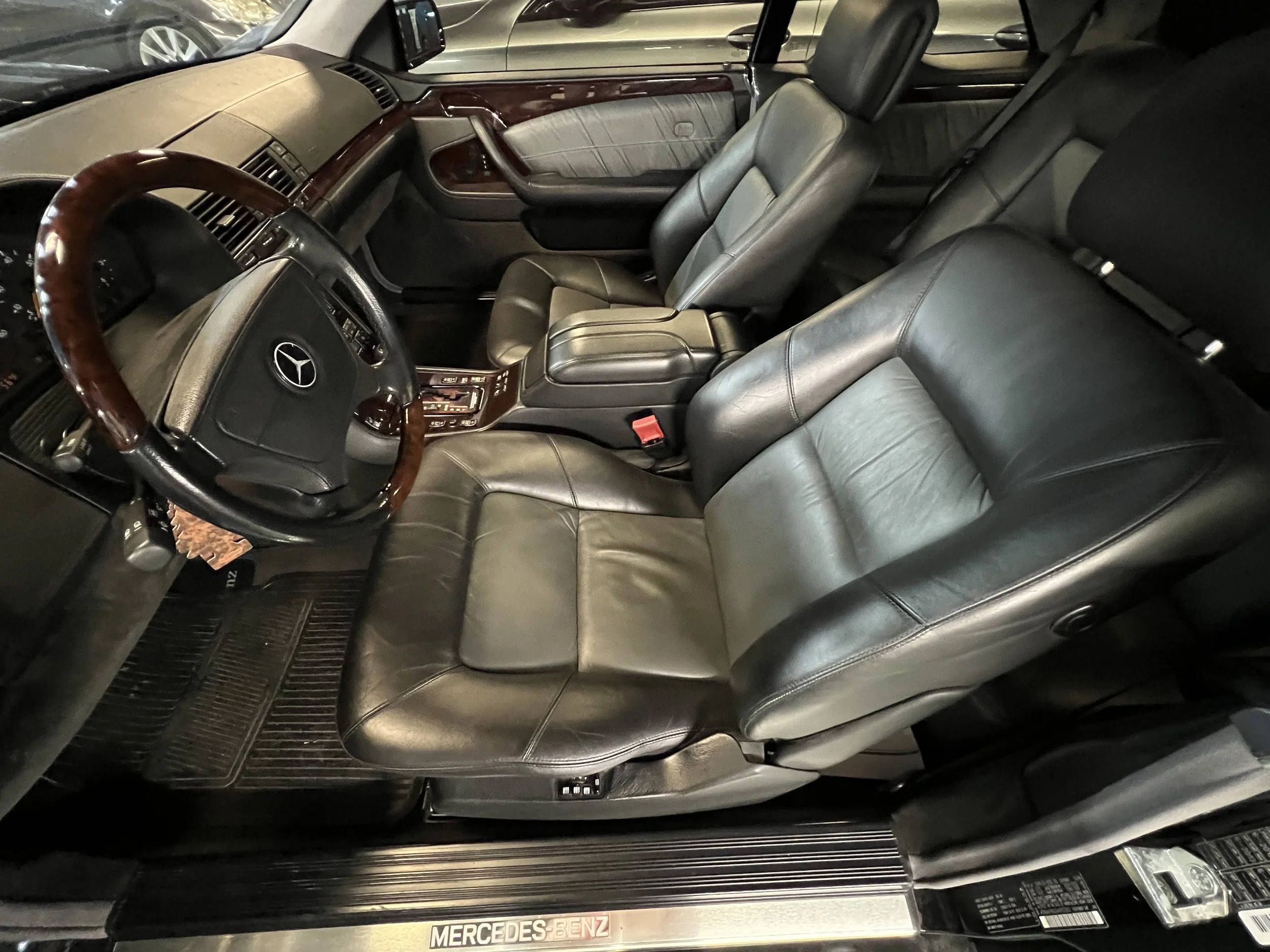 Interior of a Mercedes-Benz vehicle showing black leather seats, wood trim on the dashboard and door panels, and a steering wheel with a Mercedes-Benz emblem.