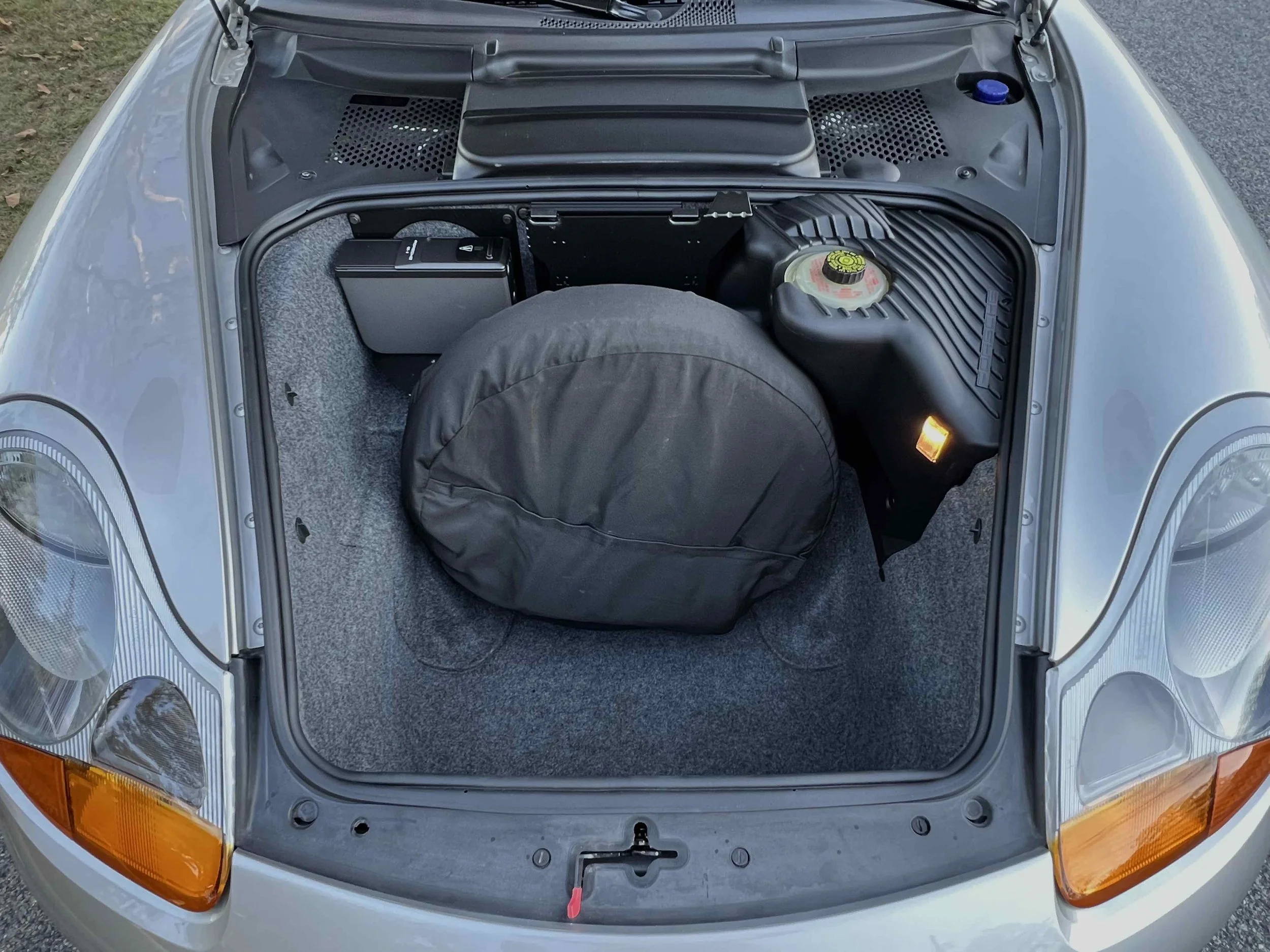 Front trunk of a silver sports car with a spare tire, a black container, and various components inside.