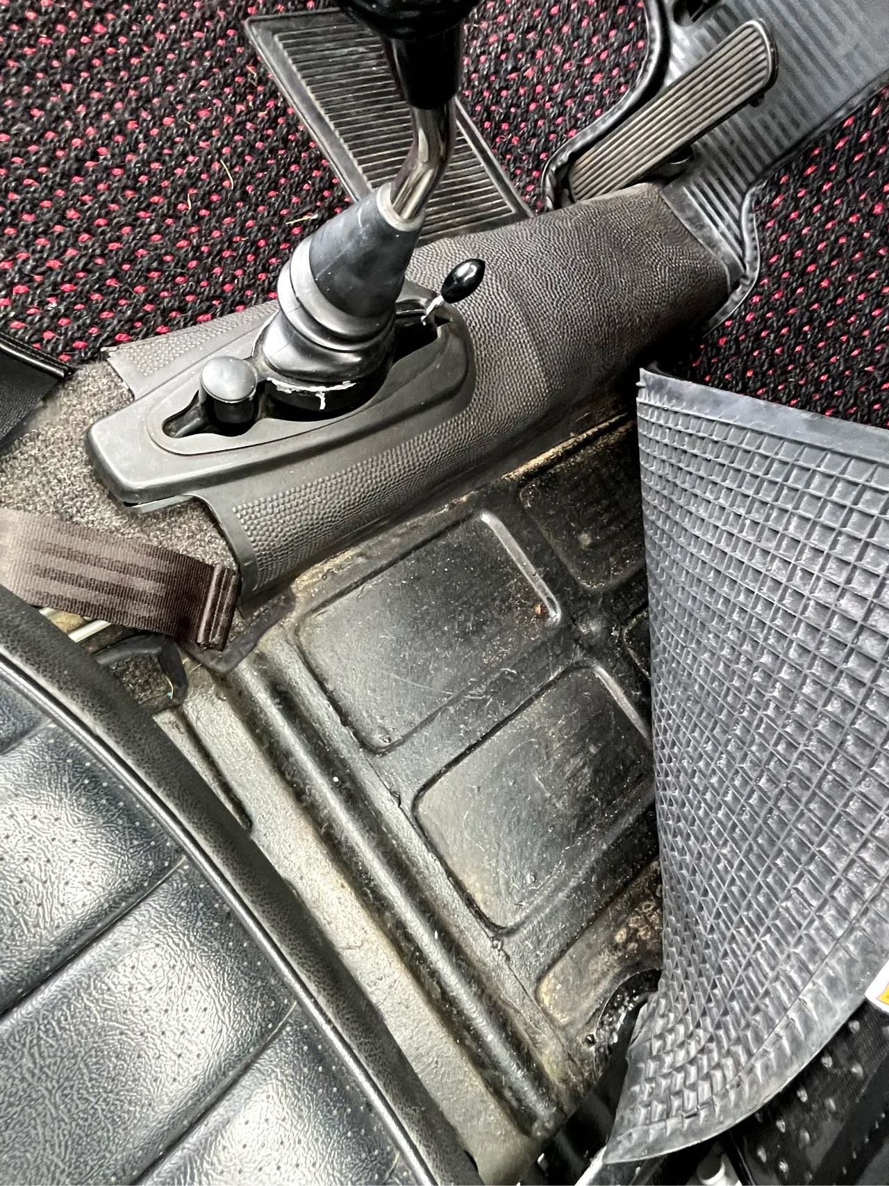 The interior of an older vehicle showing a manual gear shift, foot pedals, and a floor mat with dirt and wear.