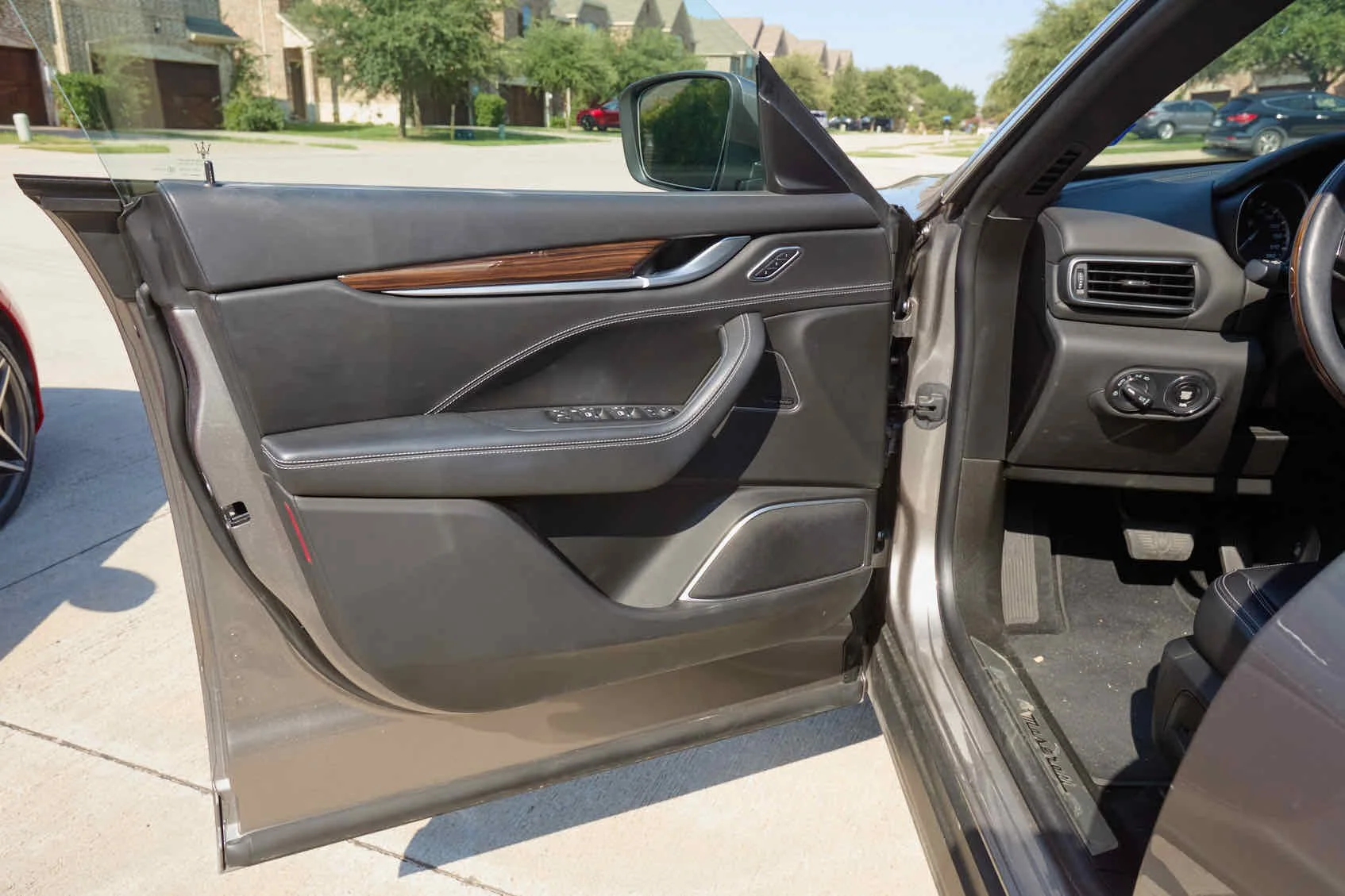The interior view of a car showing the open driver's side door, dashboard, and part of the driver's seat. The door panel is black with a wood trim accent and contains window control buttons. The dashboard is gray with a circular air vent and control 