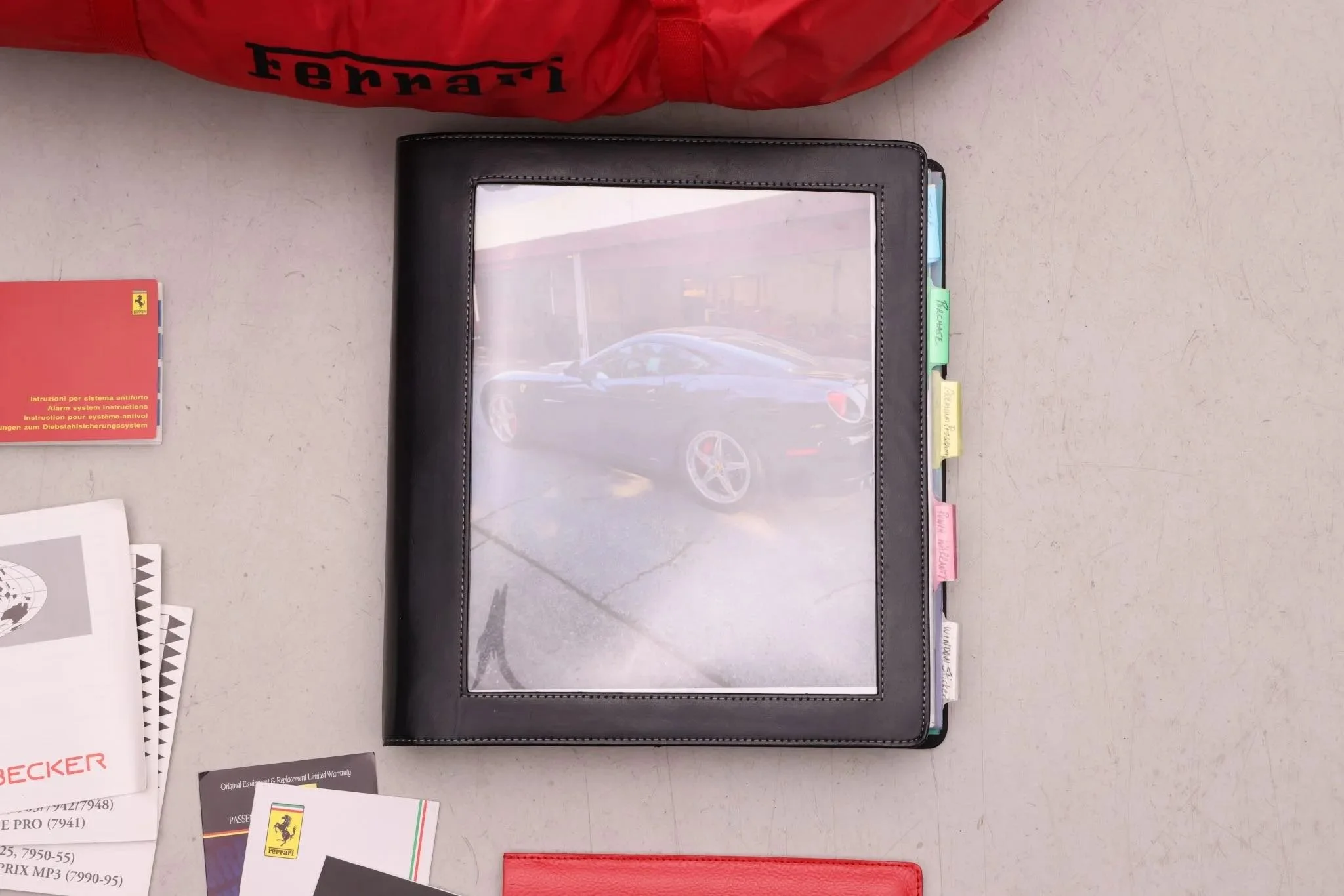 A black leather folder with colorful tabs on the right side, containing documents and a laminated photo of a black sports car. There are various papers and booklets on the table, including a red card, a Ferrari sticker, and warranty information.
