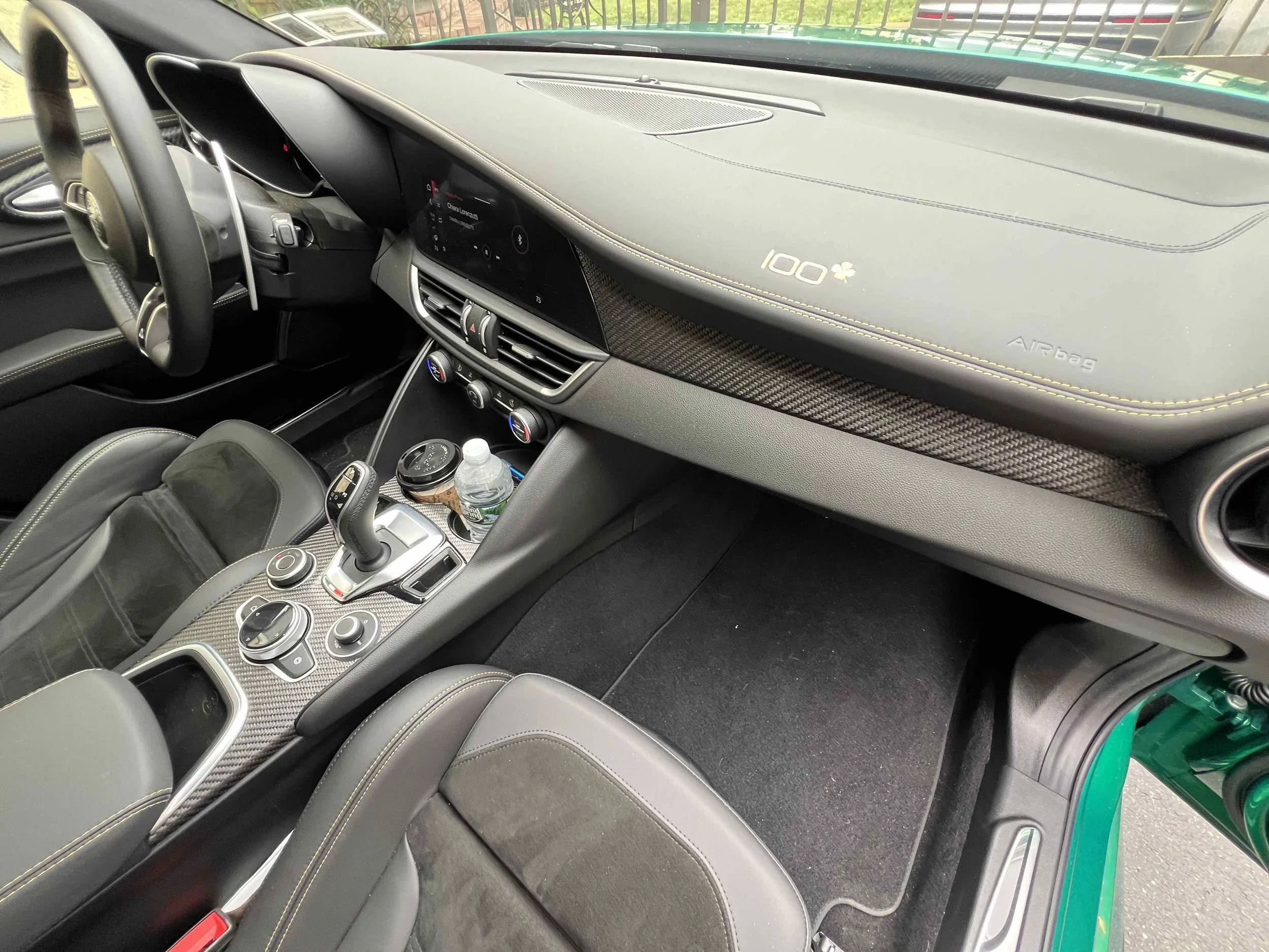 Interior of a sports car showing the dashboard, steering wheel, center console, and passenger seat. The dashboard has a digital display, air vents, and carbon fiber accents. The center console features electronic controls, a gear shifter, and a water