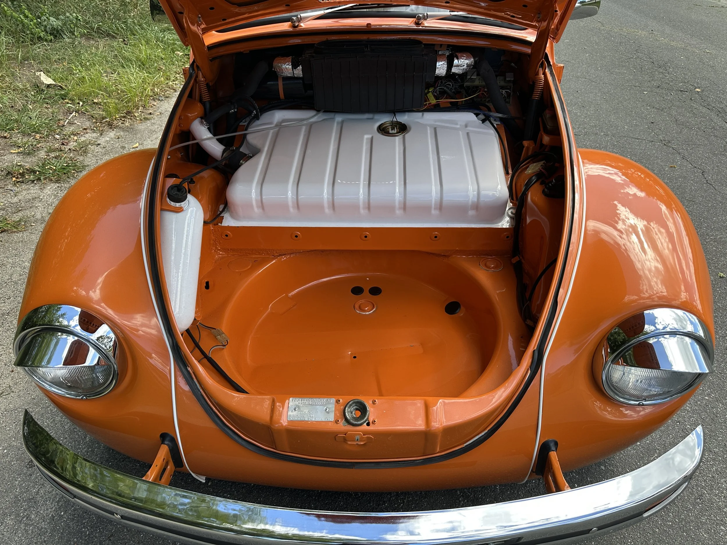 An orange vintage car with its front hood open, showing the empty engine bay with a silver-colored fuel tank and some wiring, parked outdoors on a road.