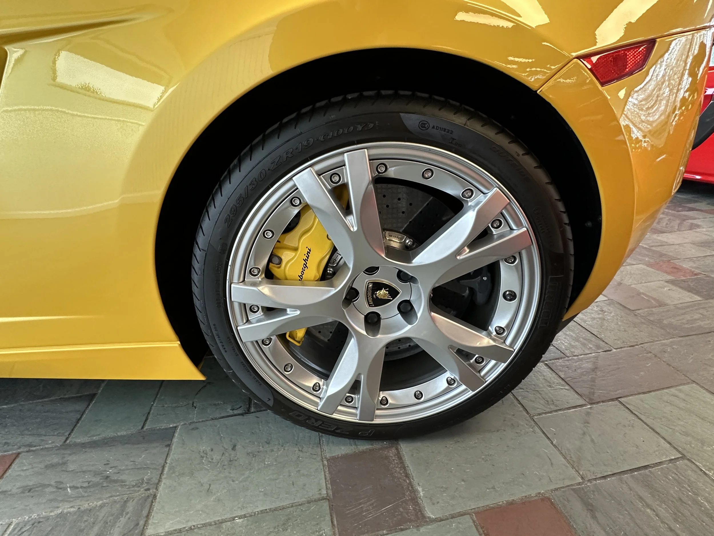 Close-up of a yellow sports car's alloy wheel, brake caliper, and tire, parked on a tiled floor.