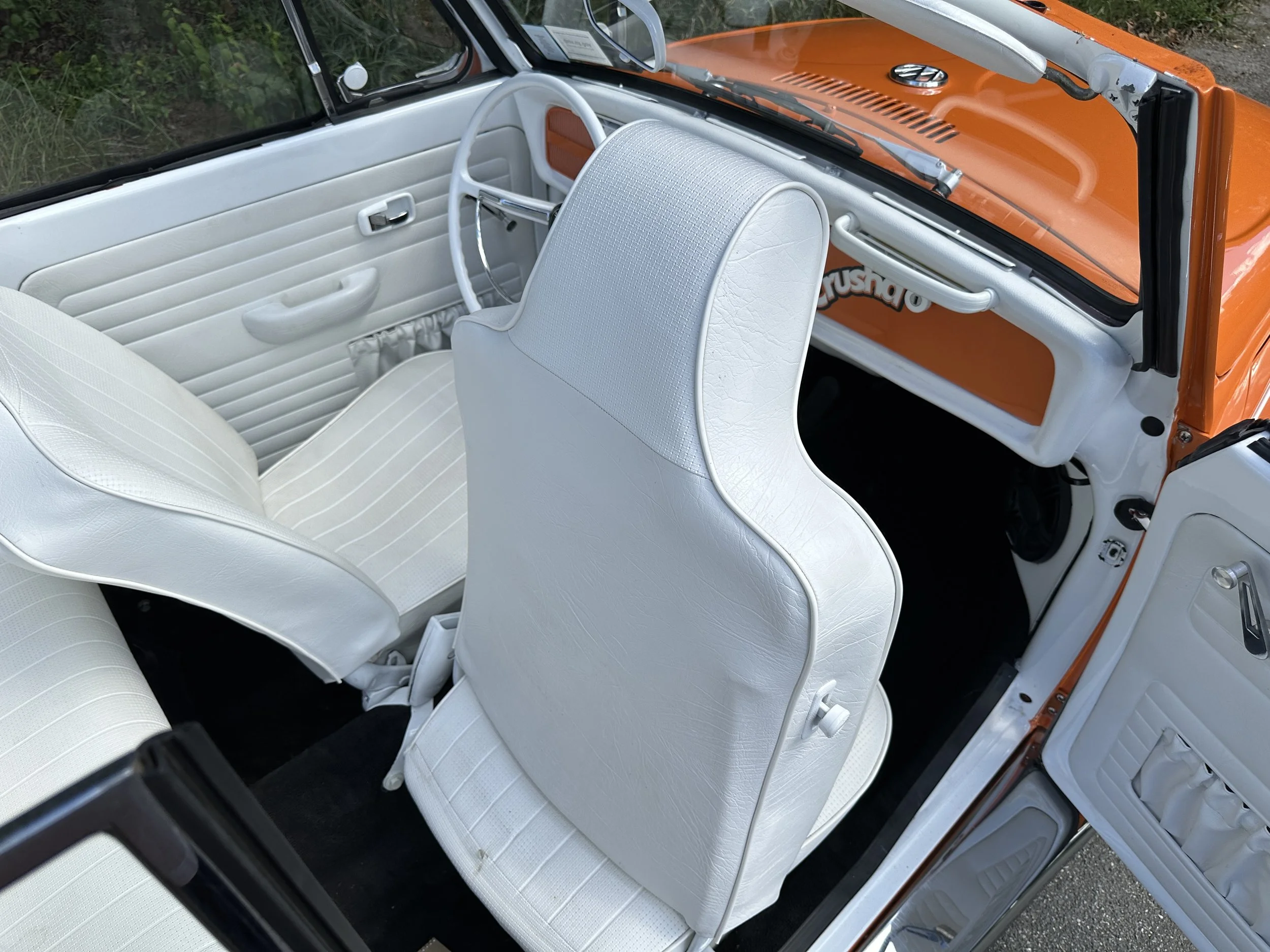 Interior of a vintage Volkswagen Beetle with white seats, white door panels, and an orange dashboard. The driver's seat and front passenger seat are visible, with the dashboard showing a steering wheel, radio, and other controls.
