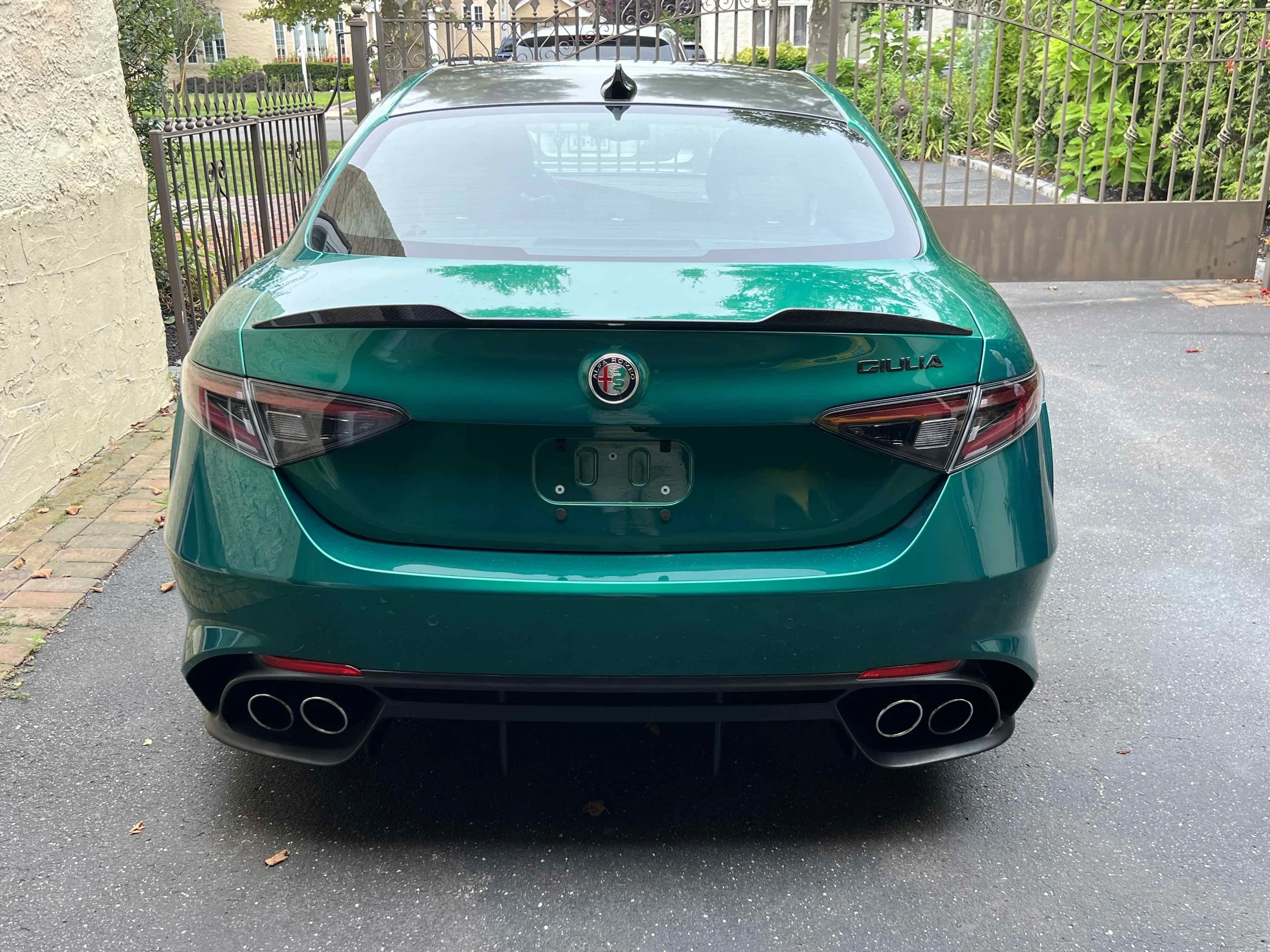 Rear view of a teal green Alfa Romeo Giulia parked on a driveway, with a closed gate and greenery in the background.