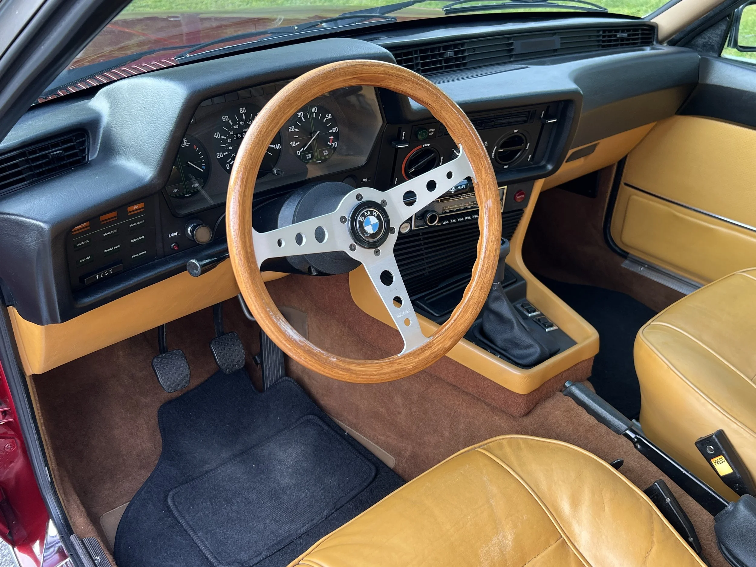 Inside the dashboard of a vintage BMW BMW 630 csi, with a wooden steering wheel, yellow seats, and black dashboard controls.