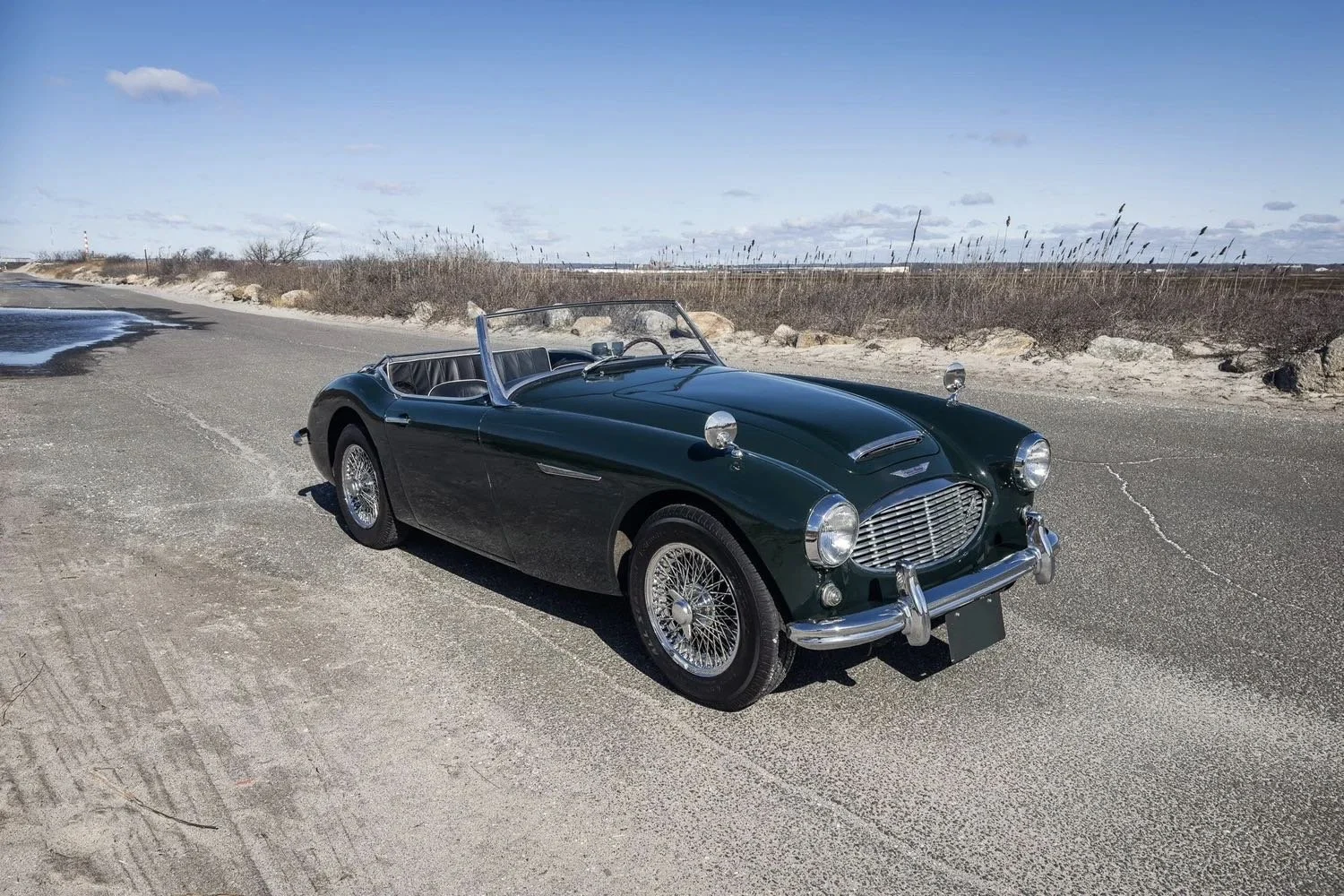 A vintage dark green convertible car parked on a sandy roadside with a clear blue sky and sparse bushes in the background.